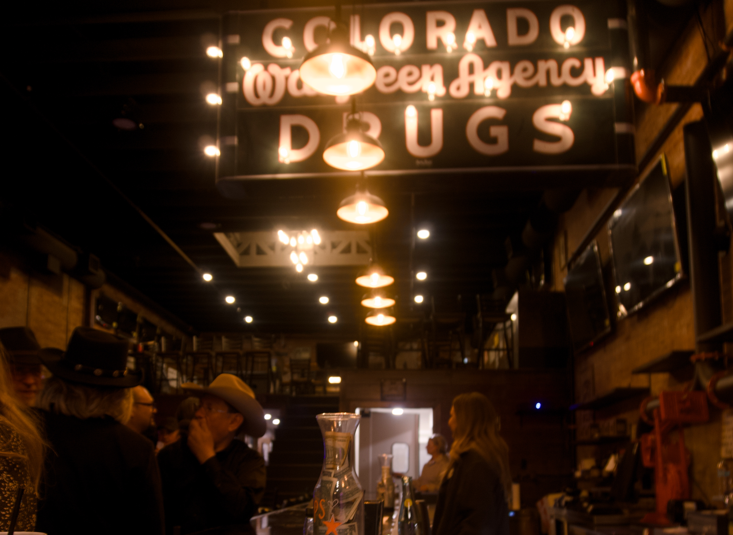 Interior of a bar or restaurant with wooden walls, hanging lights, and several people wearing cowboy hats engaged in conversation at the bar. A neon-style sign above reads 'Colorado Wat Green Agency Dugs'.