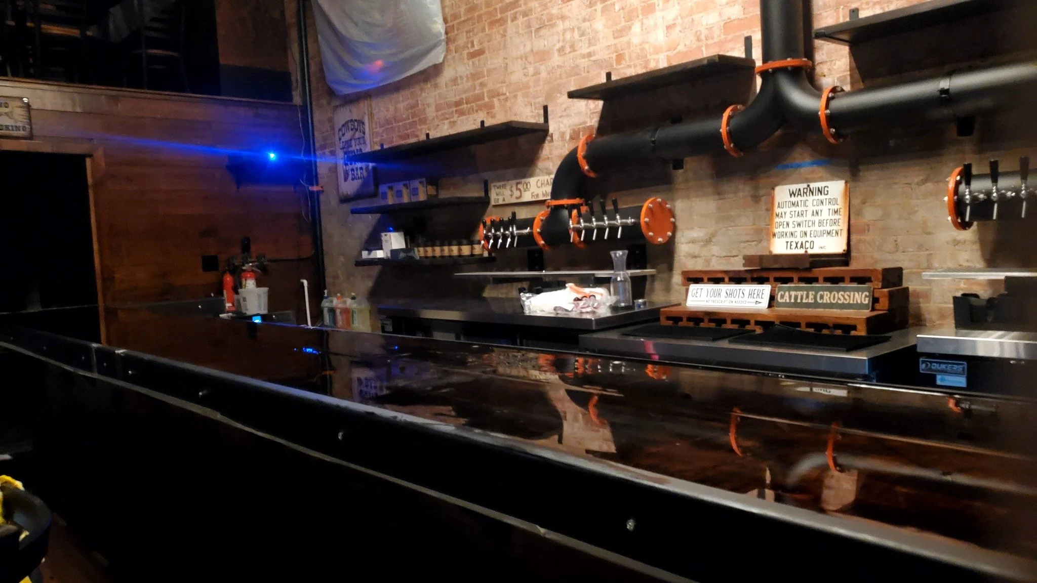 Interior of a bar or pool hall with a pool table in the foreground, brick walls, pipes, and shelves with signs and objects in the background.