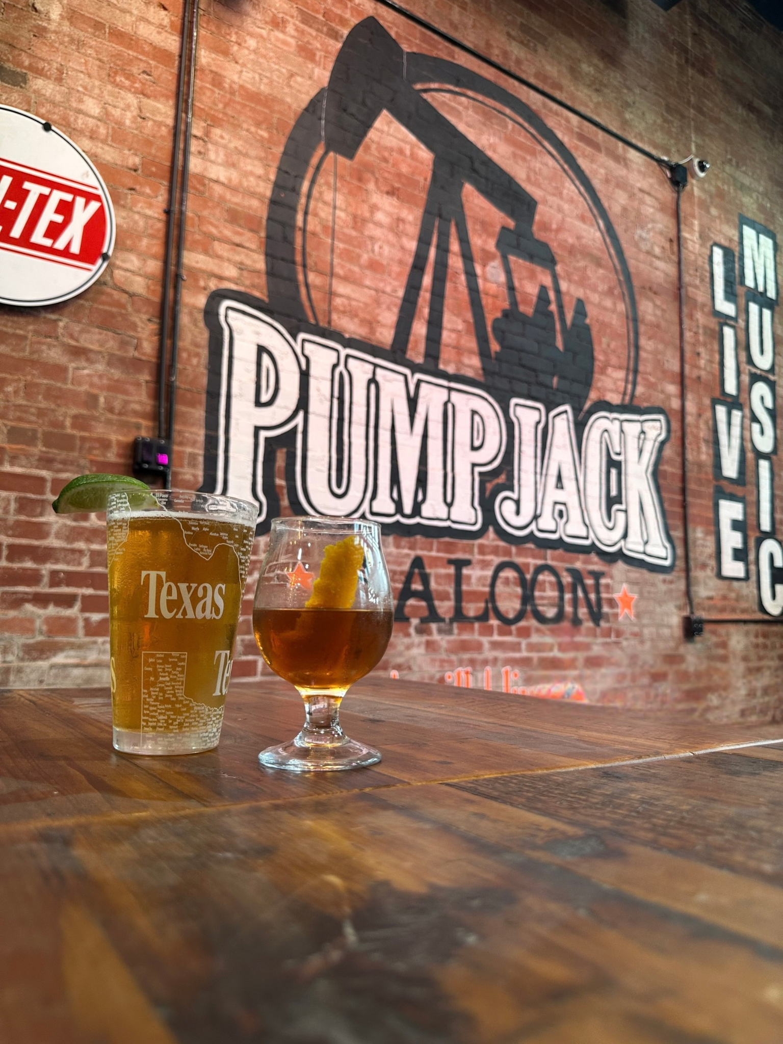 Two drinks on a wooden table in front of a brick wall with PUMPJACK TAVERN logo. One drink has a lime wedge and appears to be beer, the other has a lemon twist and looks like whiskey or bourbon.