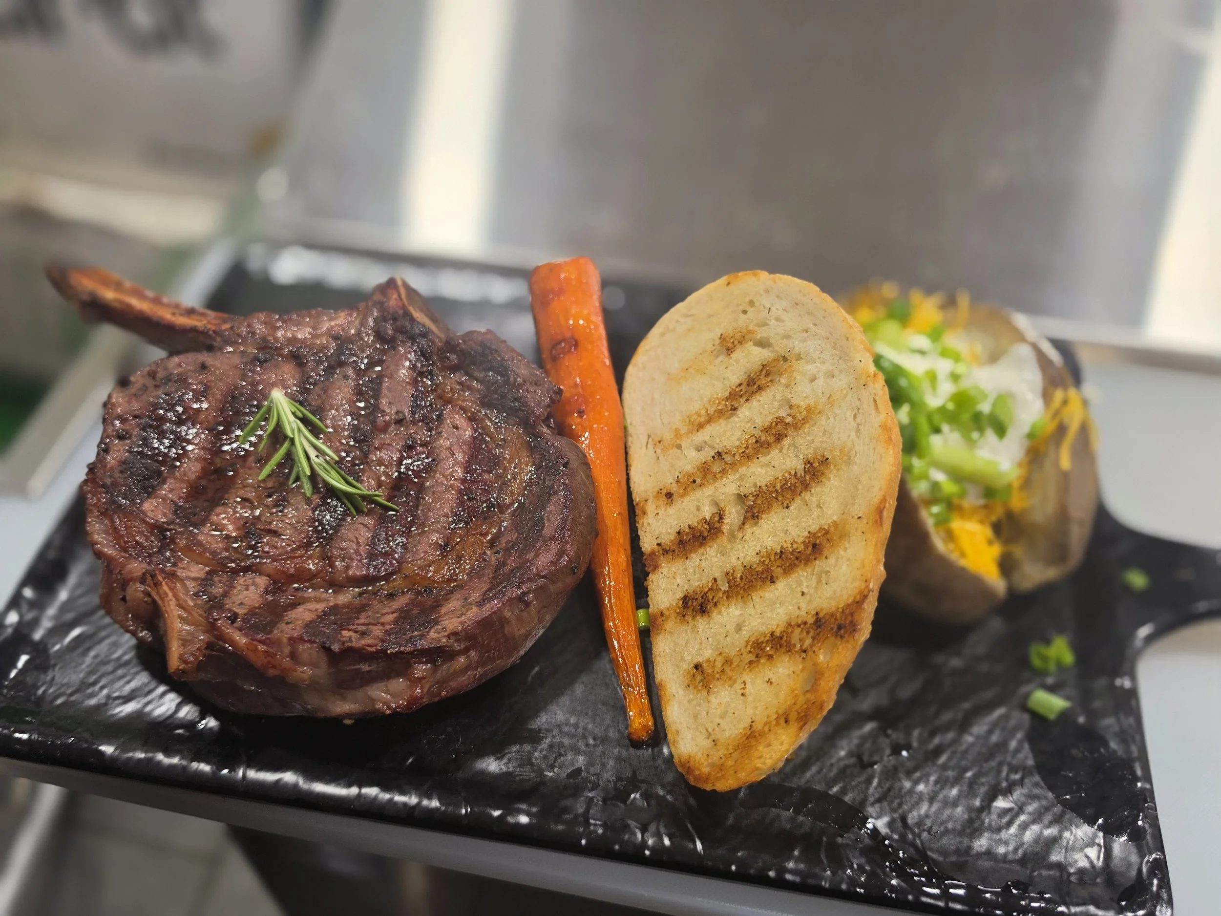 A baked bone-in steak with grill marks, garnished with a sprig of rosemary, accompanied by a roasted carrot, a toasted bread slice, and a small salad with greens, on a black serving plate.