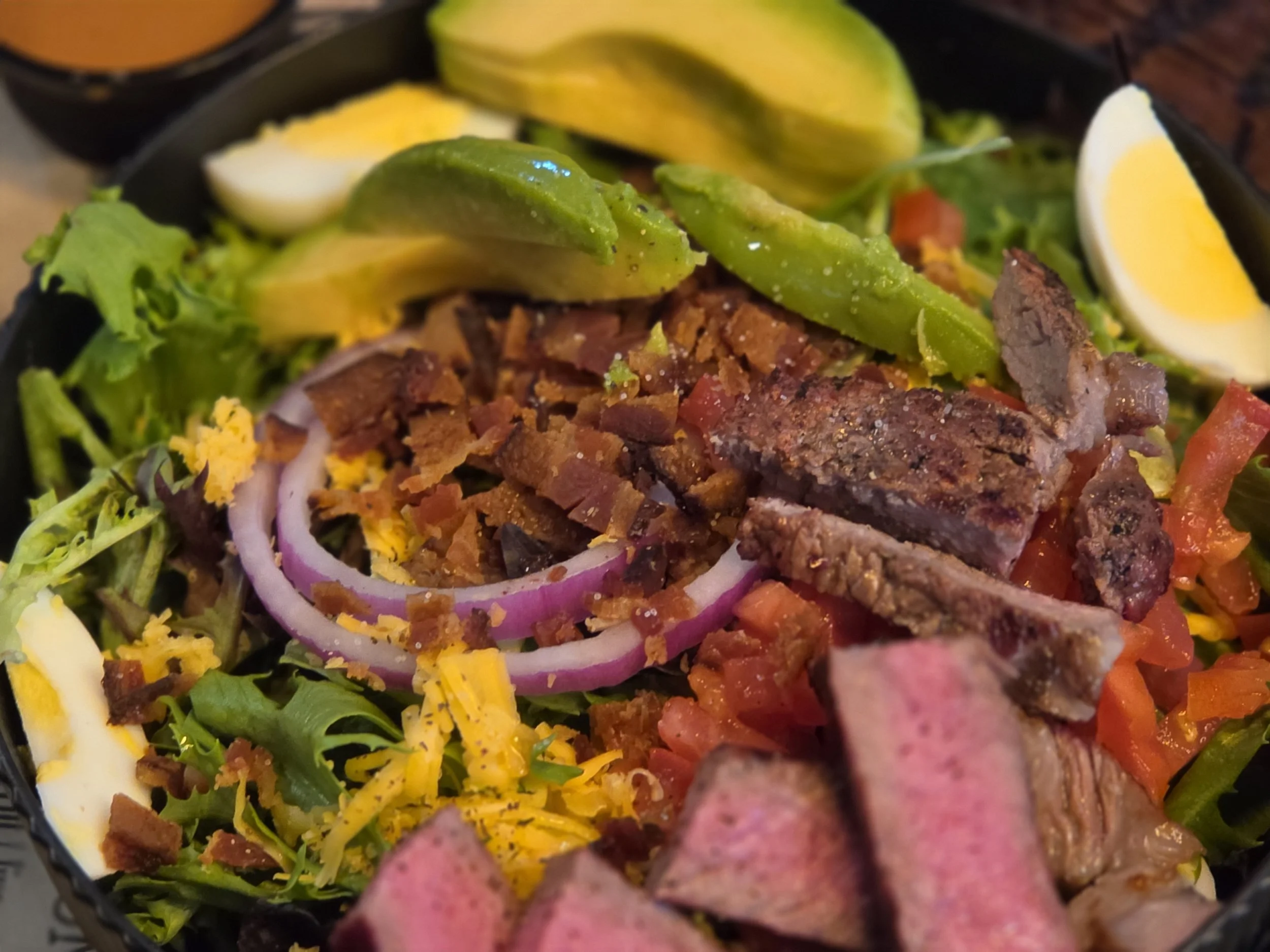 Close-up of a salad with ingredients including lettuce, sliced avocado, hard-boiled egg, sliced red onion, diced tomato, shredded cheese, and sliced cured meat, all arranged in a bowl.