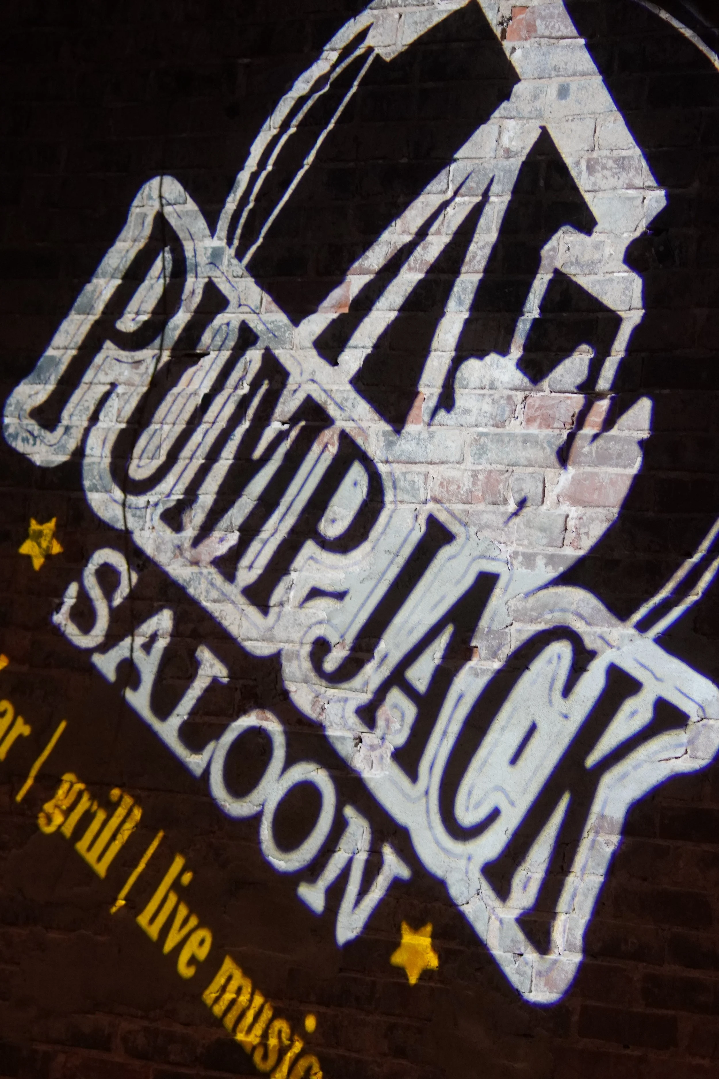 Neon sign with a white and blue outline of a check mark and a check mark in a diamond shape, with yellow stars and yellow text on a dark brick wall background.