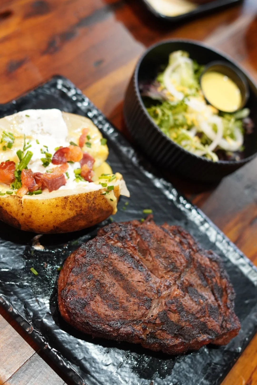 A grilled T-bone steak, loaded baked potato topped with bacon and chives, and a side salad with dressing in a black bowl.