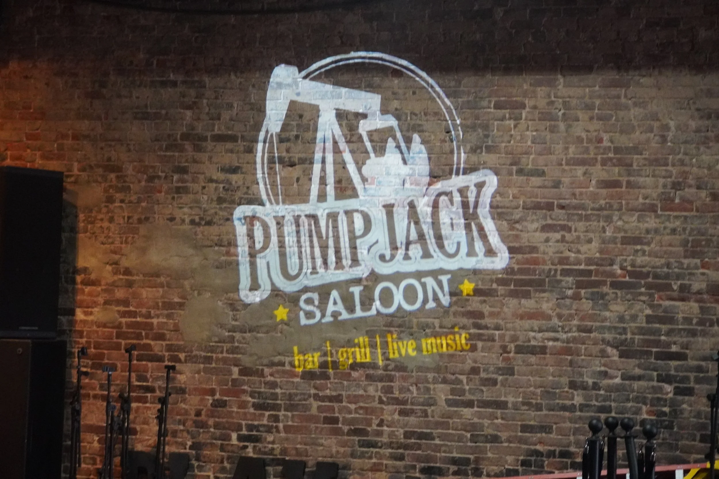 Sign on brick wall for Pumphack Saloon, featuring a circular logo with a pumpjack and stars, and text indicating it is a bar, grill, and live music venue.