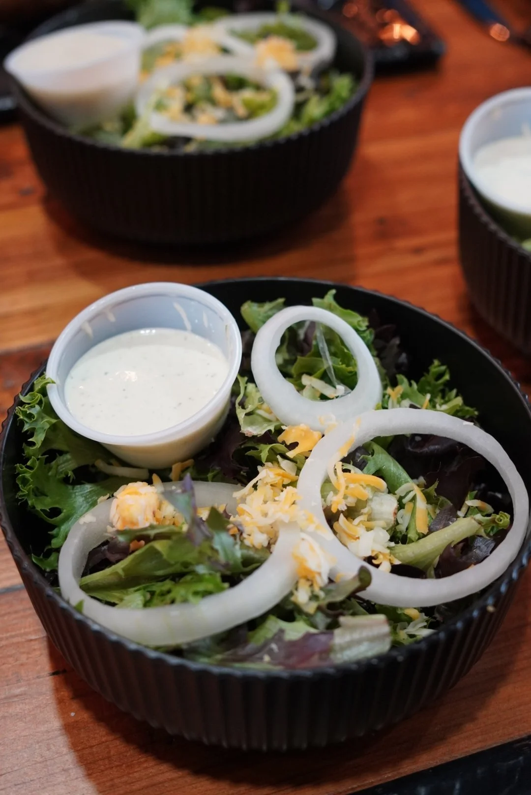 A black bowl of salad with mixed greens, sliced onions, shredded cheese, and a side of creamy dressing in a small cup, placed on a wooden table.