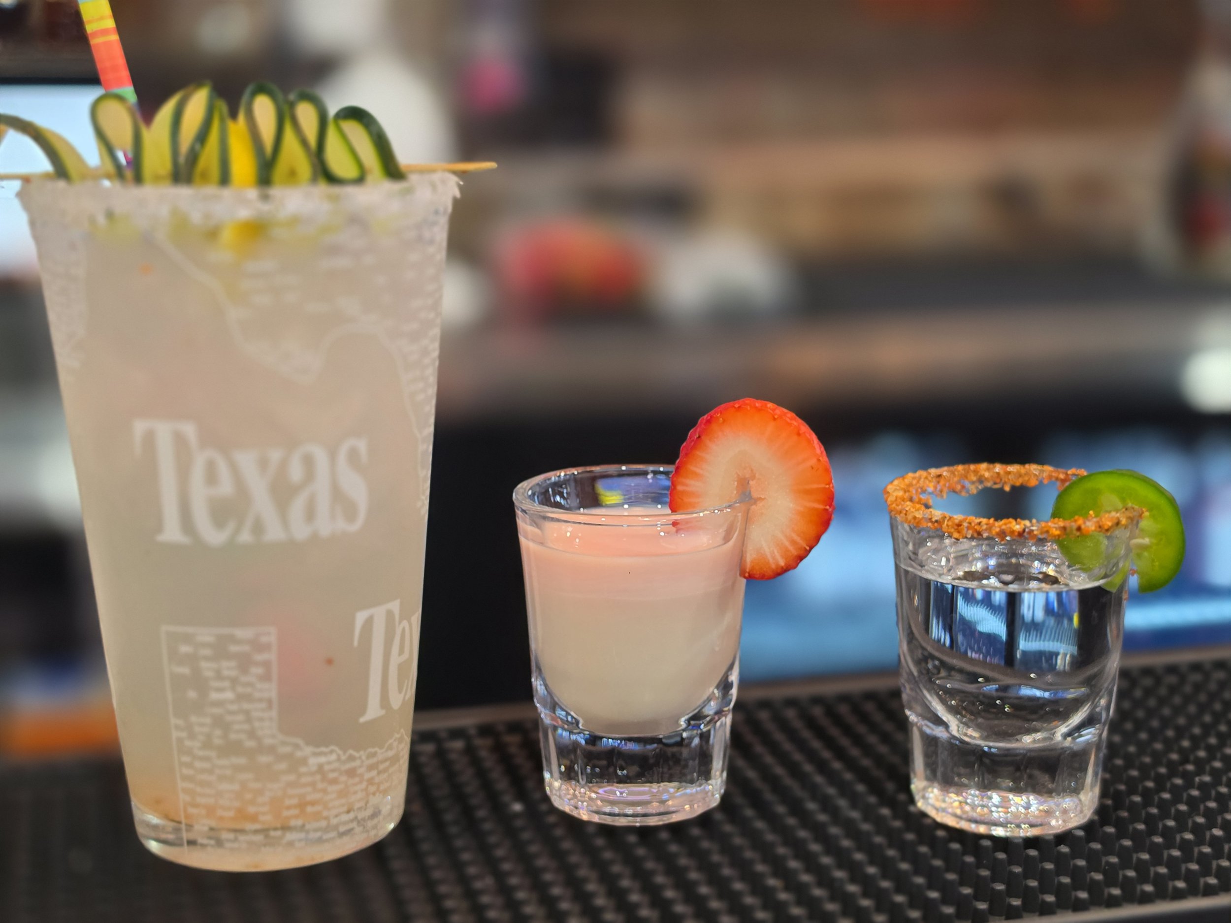 Three colorful cocktails in clear glasses with garnishes including a strawberry and a slice of lime, set on a black textured surface with a blurred background.