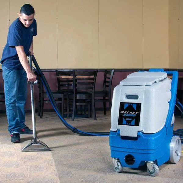 A person cleaning the carpet with a large blue and white commercial carpet cleaner in a room with chairs and a beige wall.