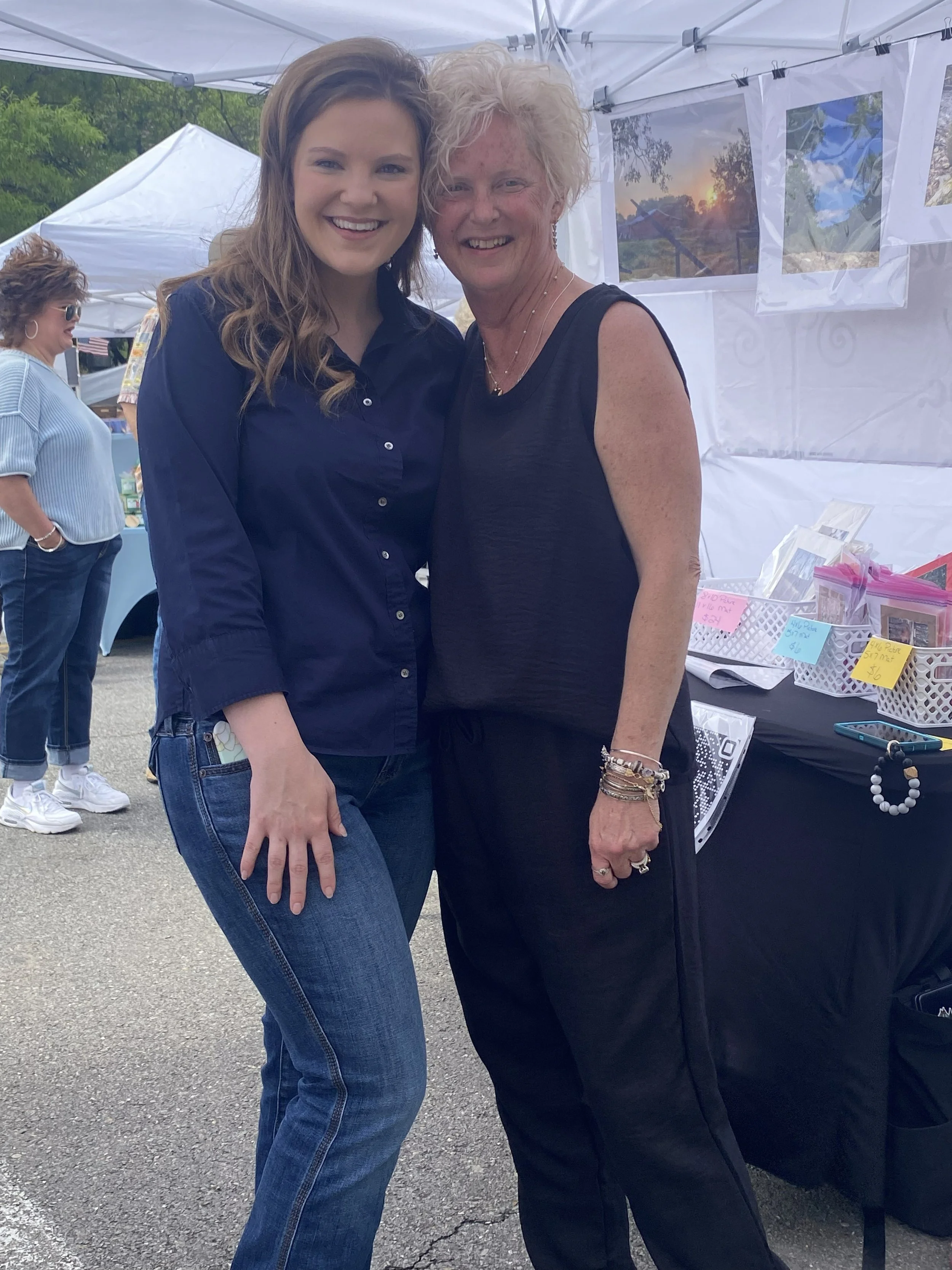 Two women smiling and standing close together outdoors at a fair or market, with tents and artwork in the background.