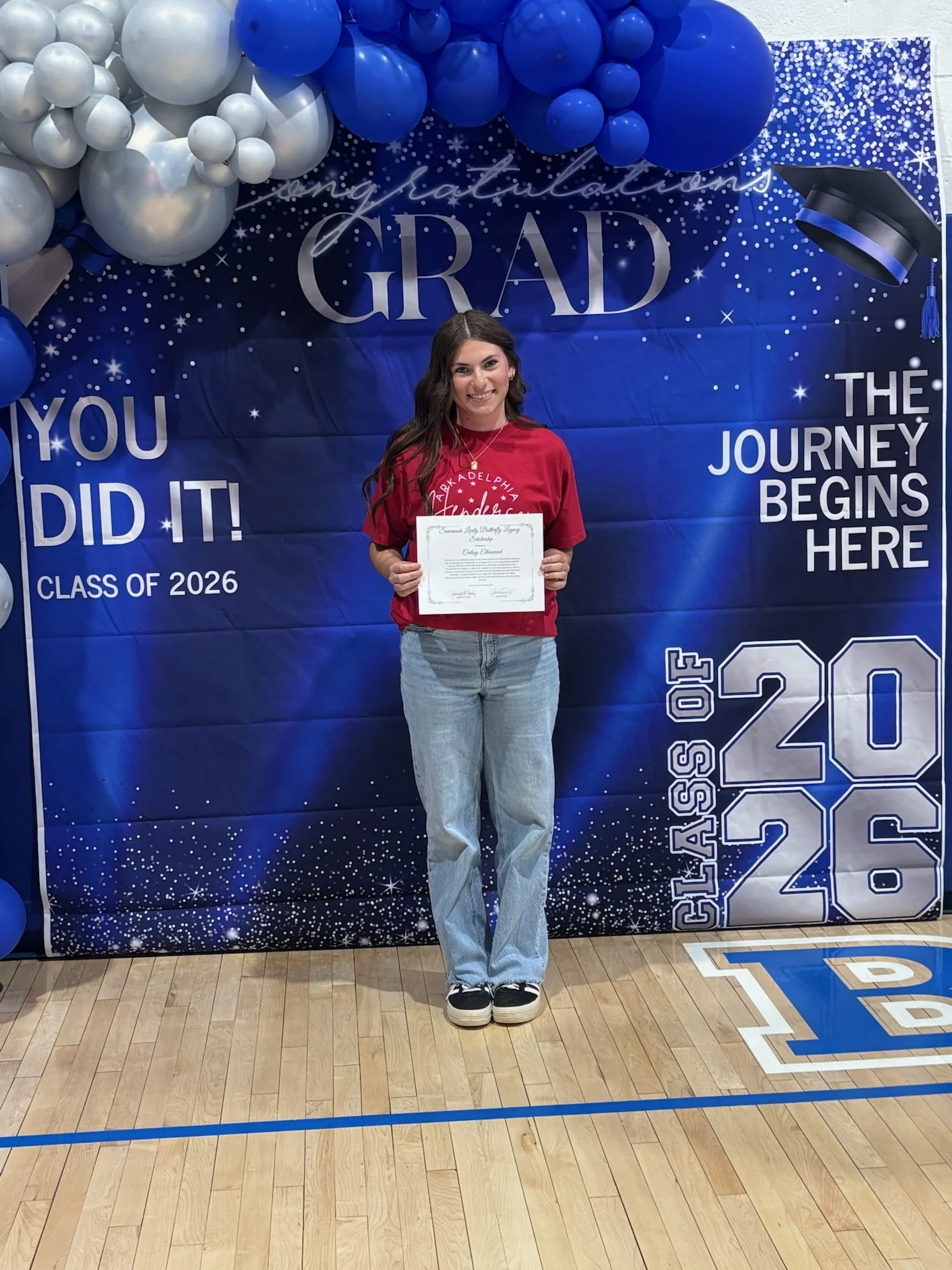 A young woman standing in front of a graduation-themed backdrop decorated with balloons and stars, holding a diploma, celebrating her graduation from high school in the class of 2026 at a basketball gym.