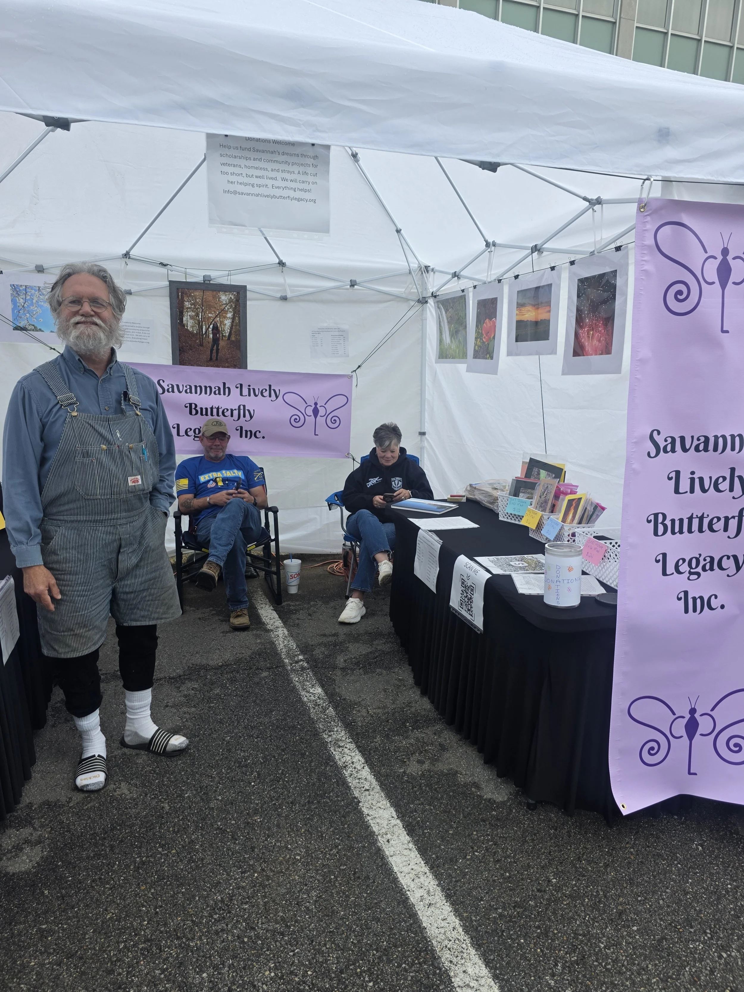 A man with gray hair and a beard standing in front of a booth at an outdoor event, with two seated people behind the booth under a white canopy. The booth displays signs and photographs related to Savannah Lively Butterfly Legacy Inc., with a purple banner and a table with books and donations container.