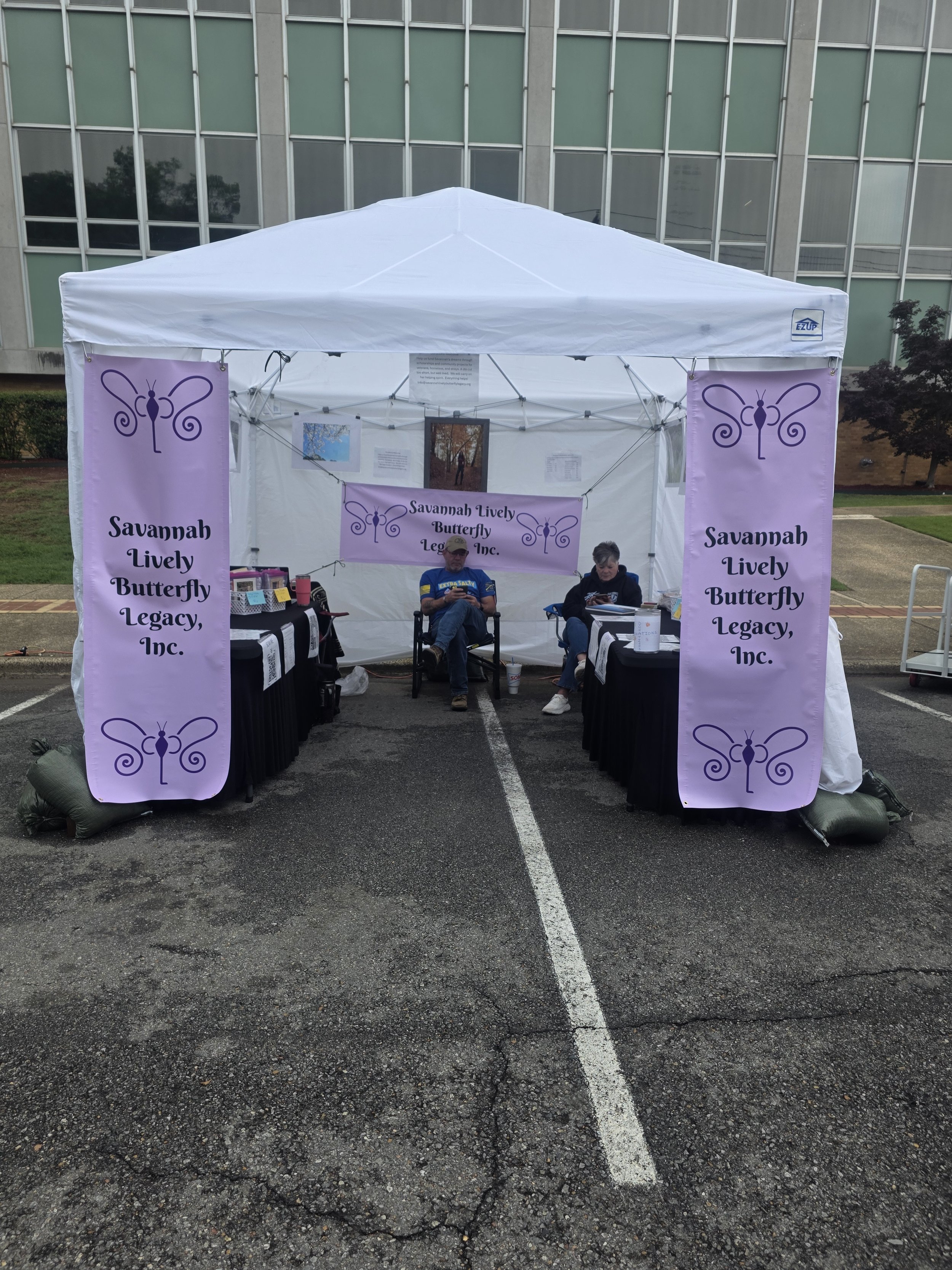 A white canopy tent set up outdoors in a parking lot, with purple banners on each side displaying a butterfly logo and the text 'Savannah Lively Butterfly Legacy, Inc.' Two people are seated inside the tent, one wearing a cap and blue shirt, the other a woman with gray hair, with tables and informational materials inside.