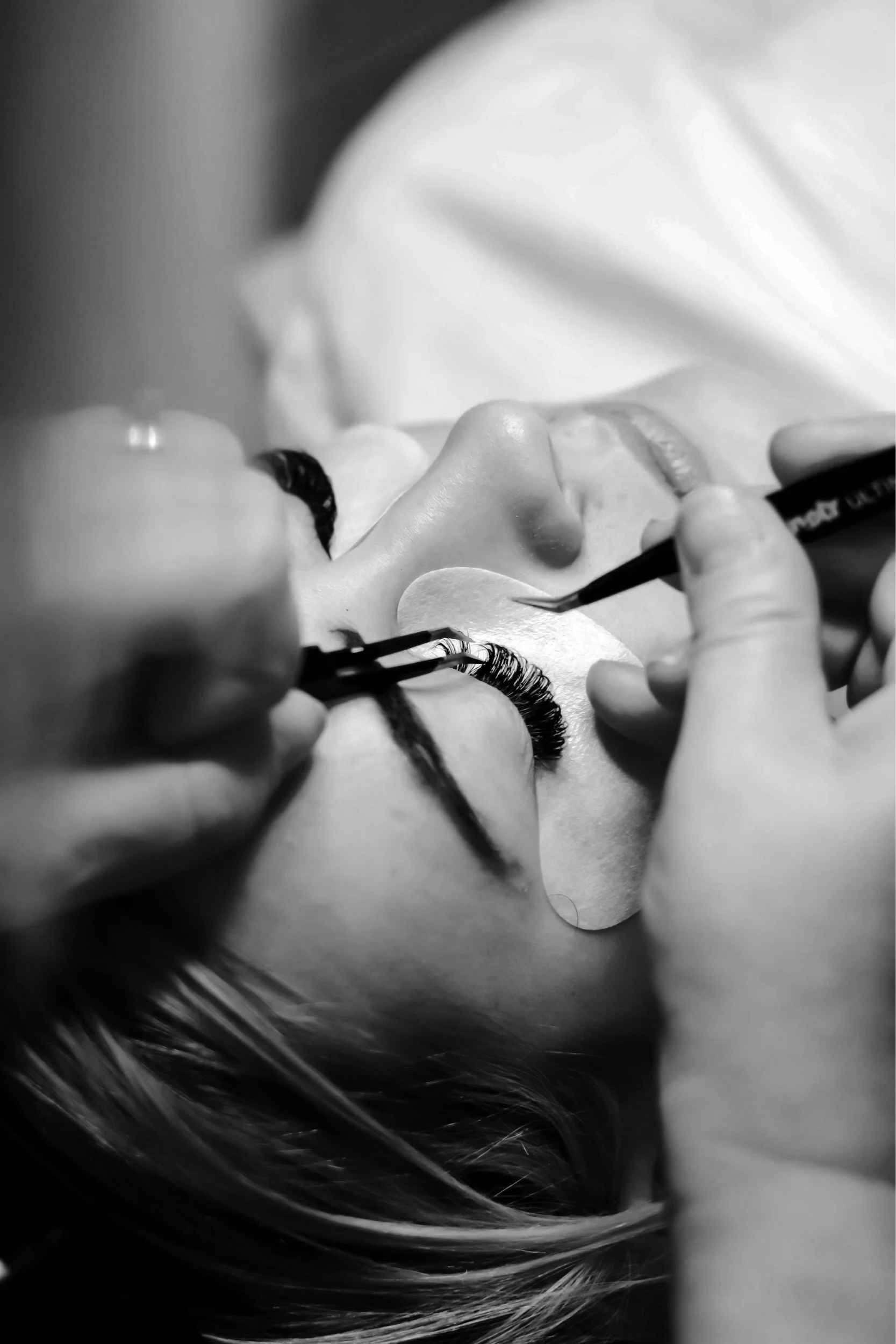 Close-up of a person receiving eyelash extensions, lying down with eyes closed, as a technician applies the false eyelashes using tweezers, in black and white.