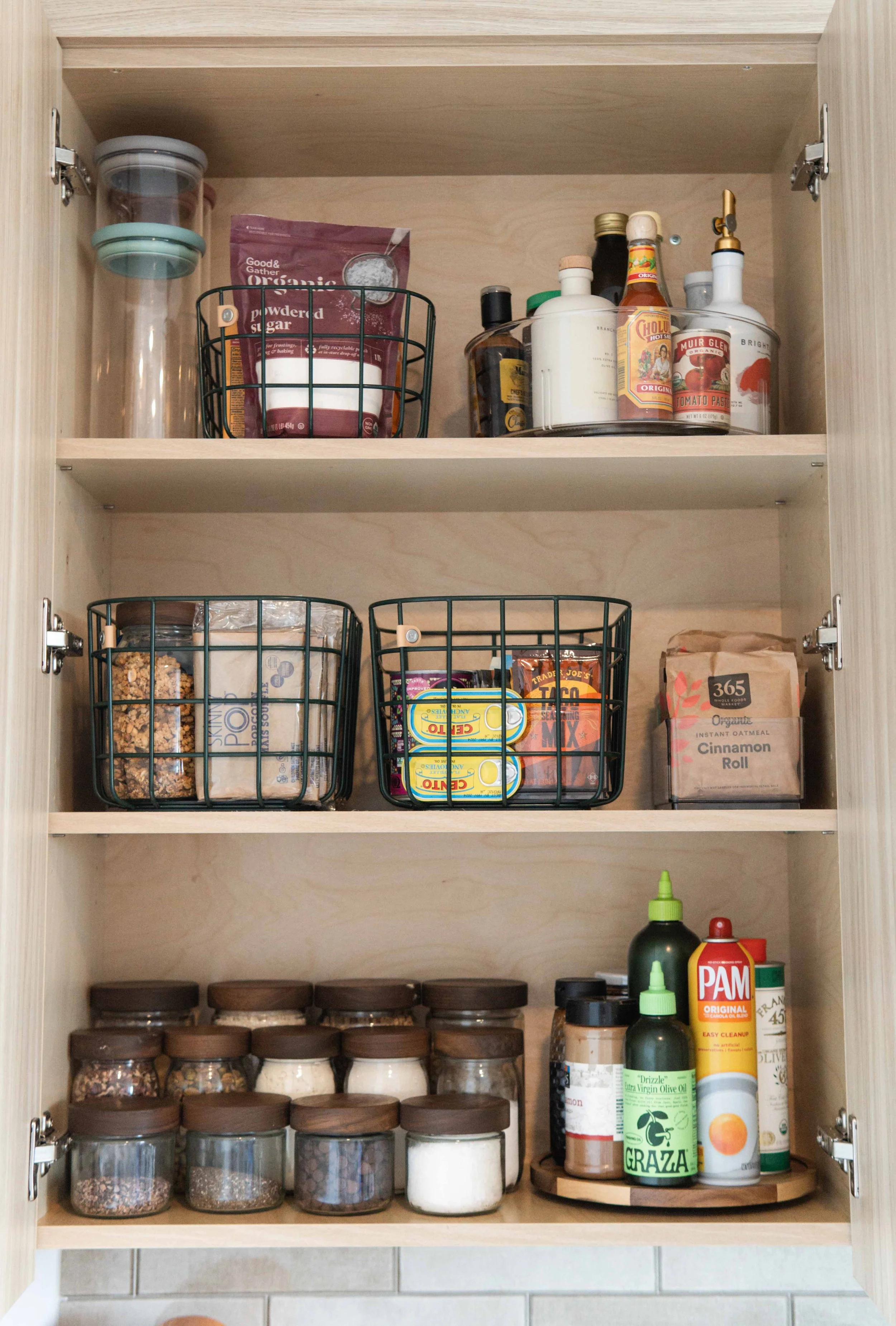 Spacious kitchen cabinet with three shelves holding jars, spices, canned goods, and condiments, including ketchup, mustard, olive oil, and instant oatmeal.