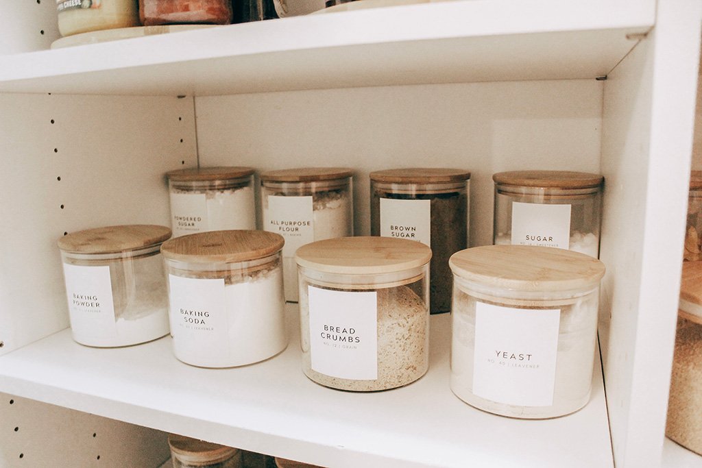 Baking ingredients in glass jars with bamboo lids on a white shelf, including flour, sugar, baking powder, baking soda, bread crumbs, yeast, and all-purpose flour.