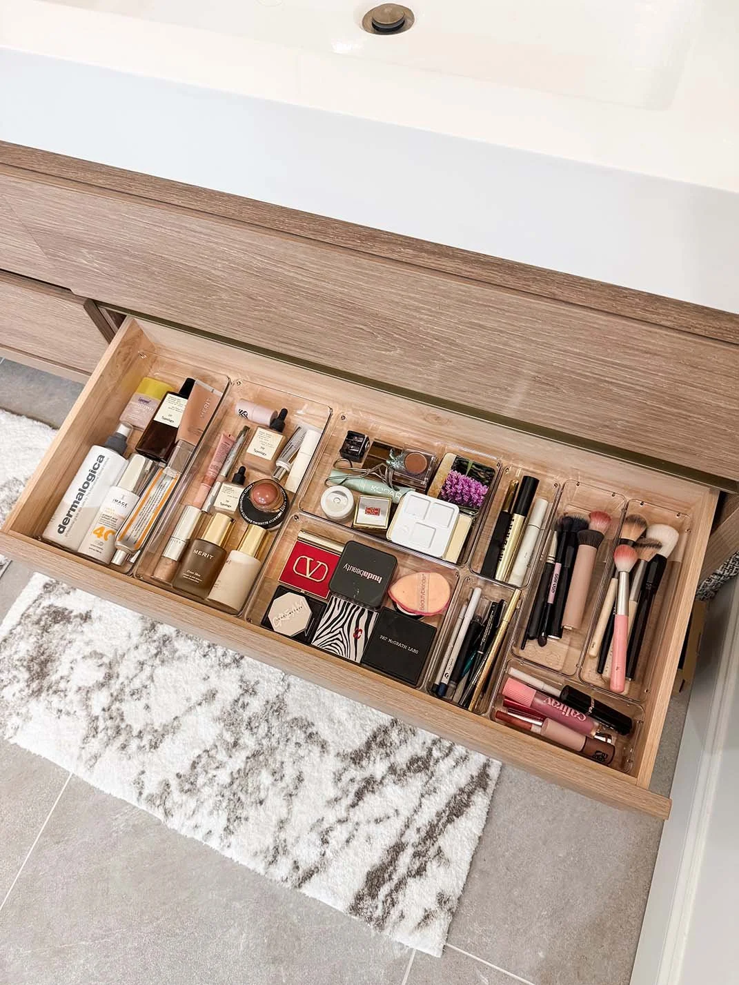 Open wooden drawer containing various makeup and skincare products, brushes, and compacts, situated in a bathroom with a white rug and gray tiled floor.