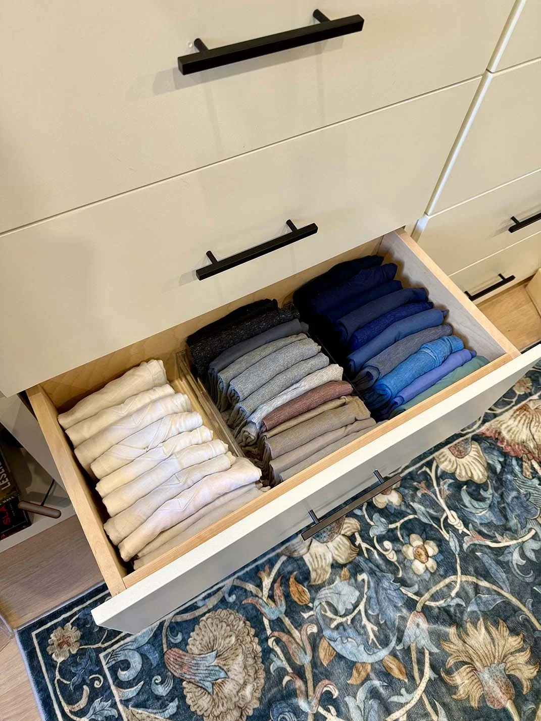 Open drawer with neatly folded socks in various colors, including white, gray, black, navy, and brown, beside a cream-colored dresser with black handles, on a patterned rug.