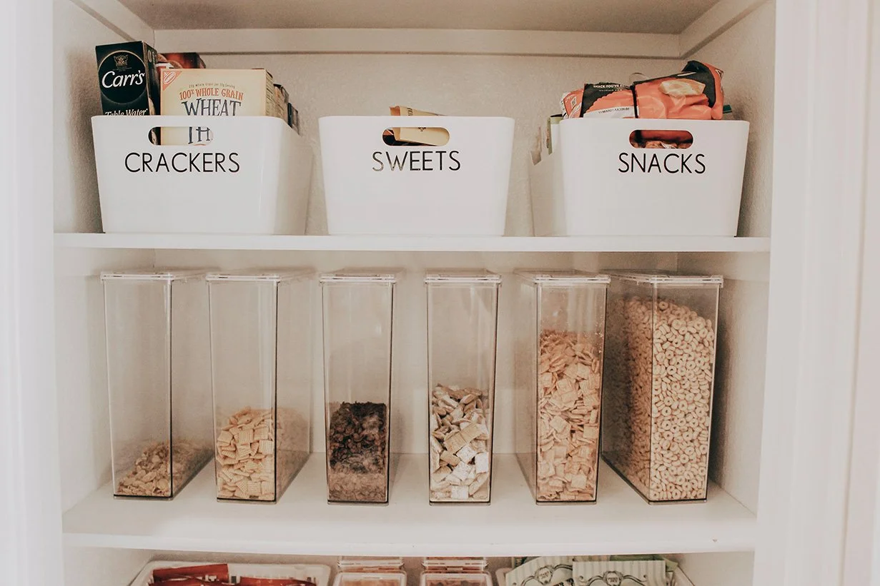 Kitchen pantry with white labeled bins for crackers, sweets, and snacks on the top shelf; clear containers of pastas, cereals, and grains on the middle shelf.