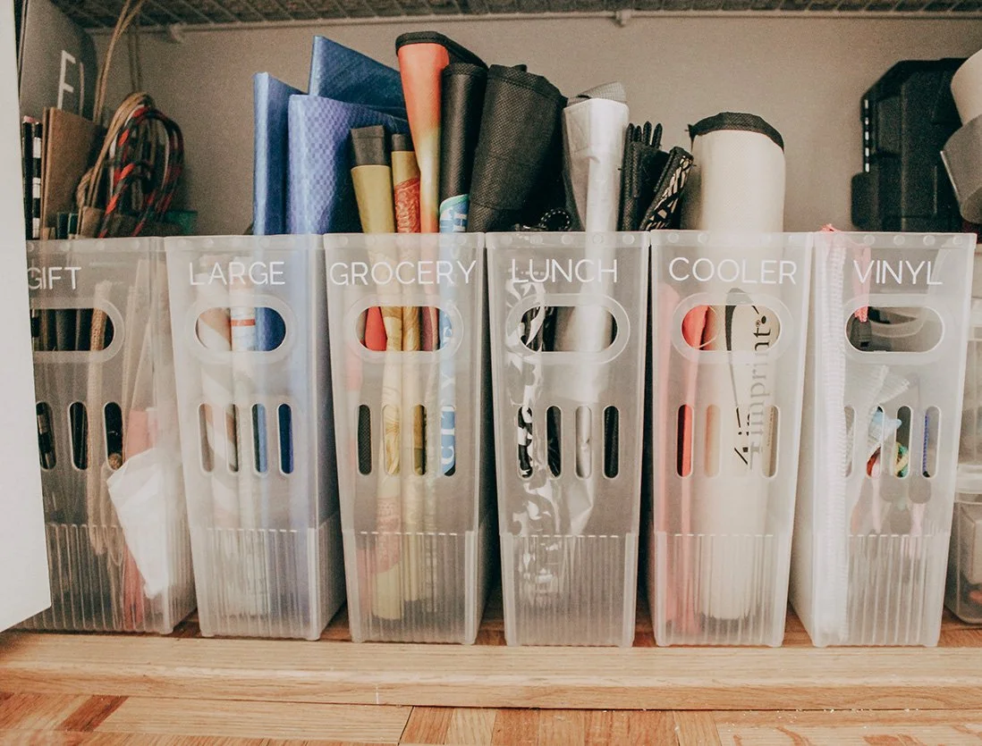Six labeled plastic storage bins containing art supplies, organized as GIFT, LARGE, GROCERY, LUNCH, COOLER, VINYL, on a wooden shelf.
