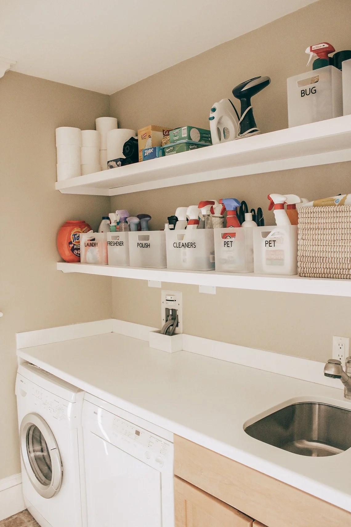 Laundry room with shelves holding paper towels, cleaning supplies, and labeled containers for laundry, detergent, polish, cleaners, and pet grooming products, washing machine, and a laundry sink.