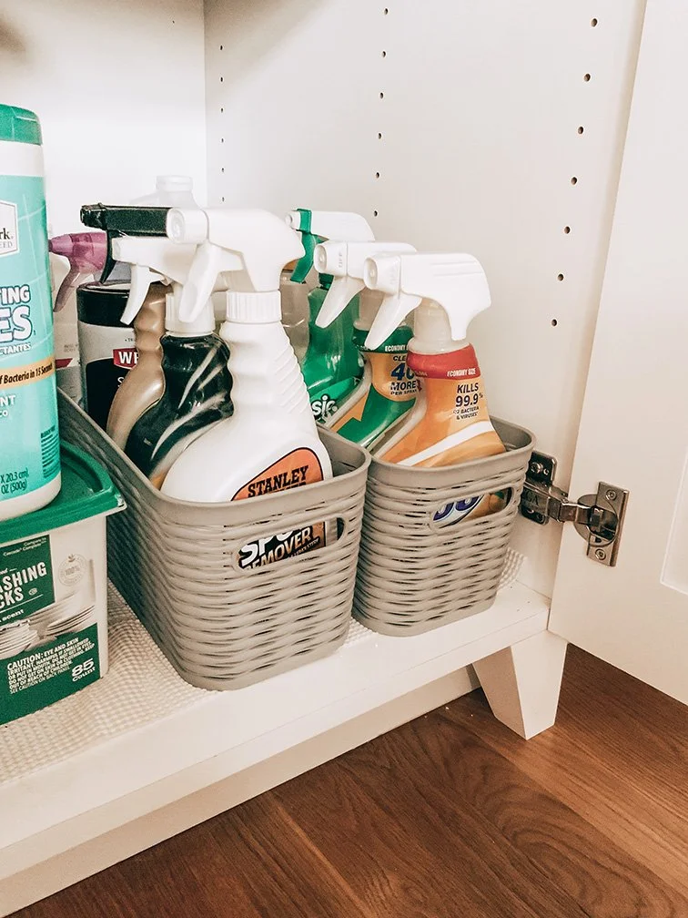 Two gray storage baskets filled with various spray bottles of cleaning products placed on a white shelf inside a cabinet.