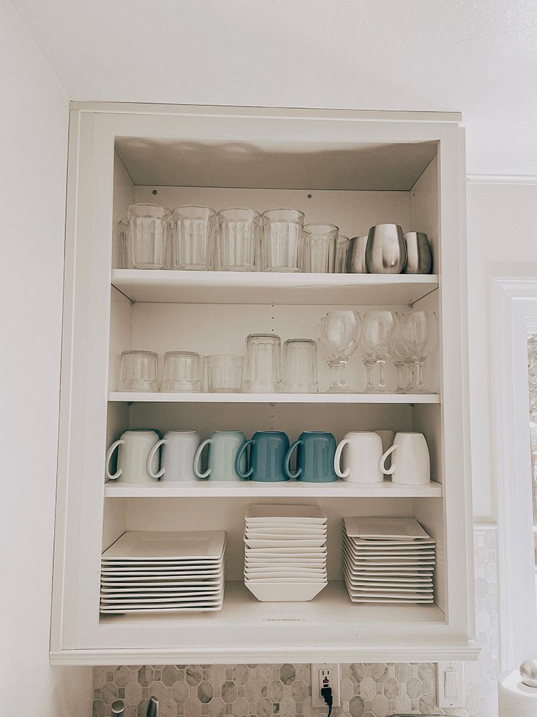 Kitchen cabinet with clear glassware, white and blue mugs, and white and blue plates inside.