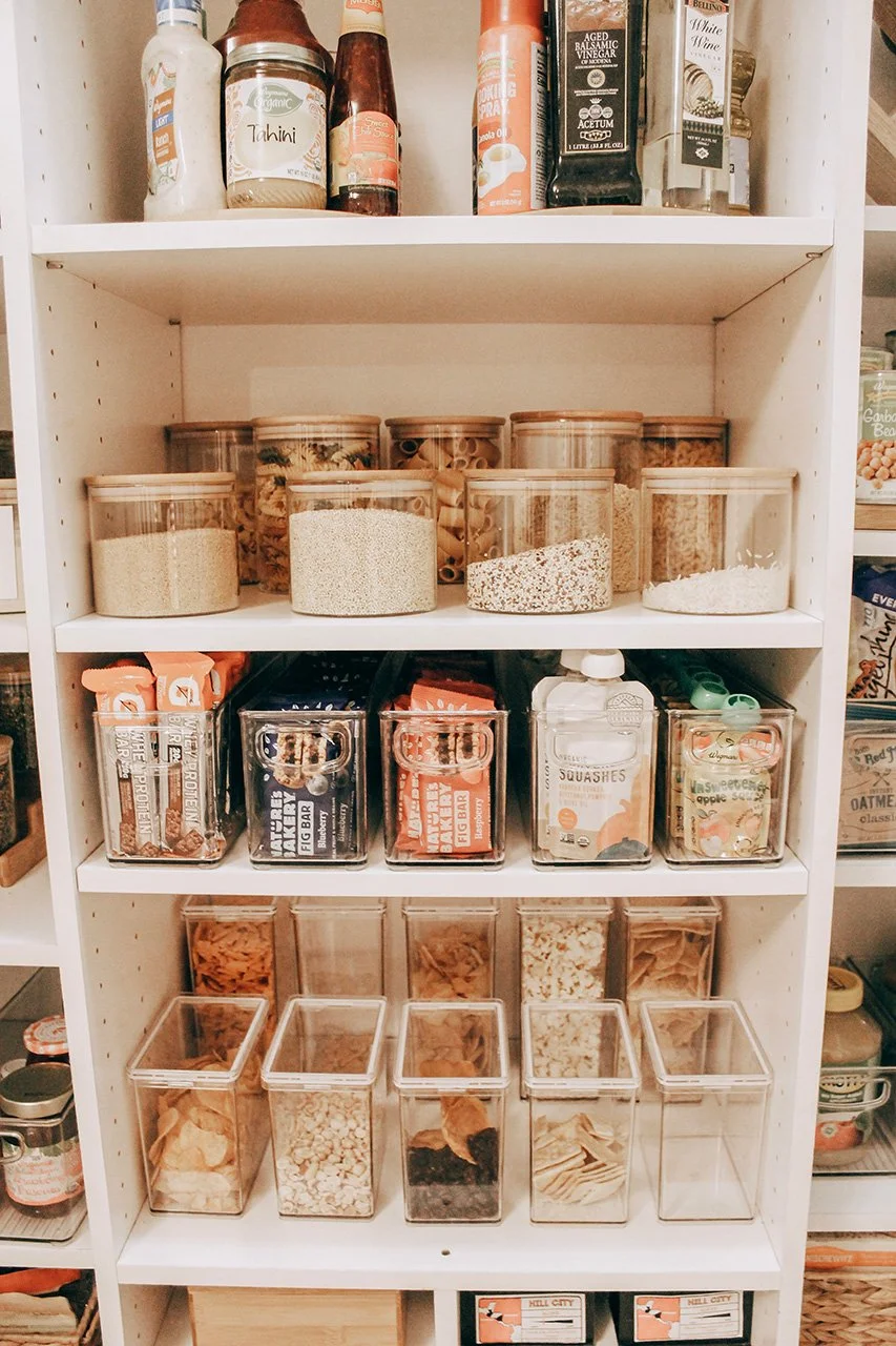White pantry shelf containing various containers of dry foods, snacks, and cooking ingredients. The top shelf has bottles and jars, the second shelf has labeled spice jars, the third shelf has boxed snack bars and a bottle of squashes, the fourth shelf contains clear plastic bins with different types of dried pasta and snacks, and the bottom shelf has more containers of dry foods.