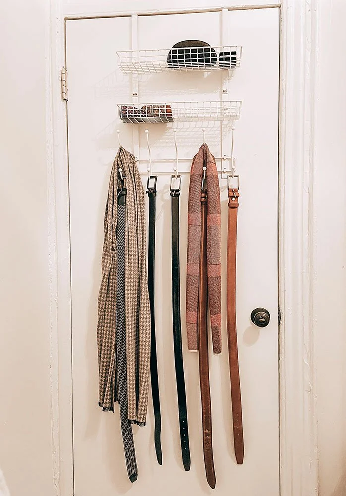 Four belts and one scarf hanging on hooks on a white door, with a white wire organizer on top holding a black hat and two small containers.