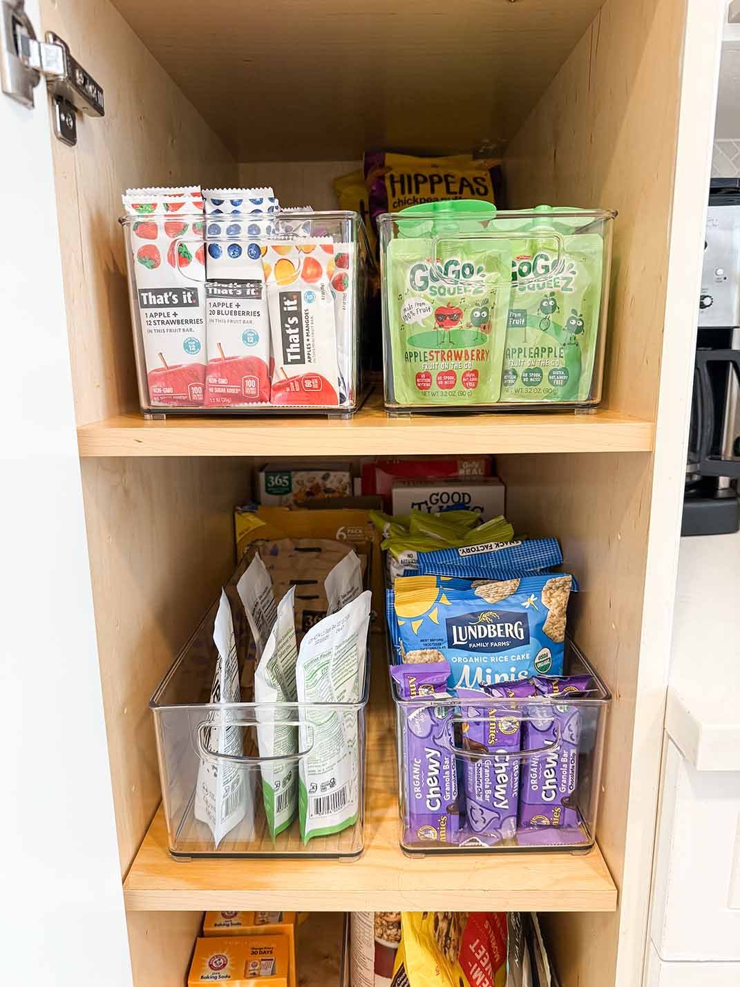 Organized pantry shelf filled with various snack packages, including fruit snacks, rice cakes, and granola bars, in clear plastic bins.