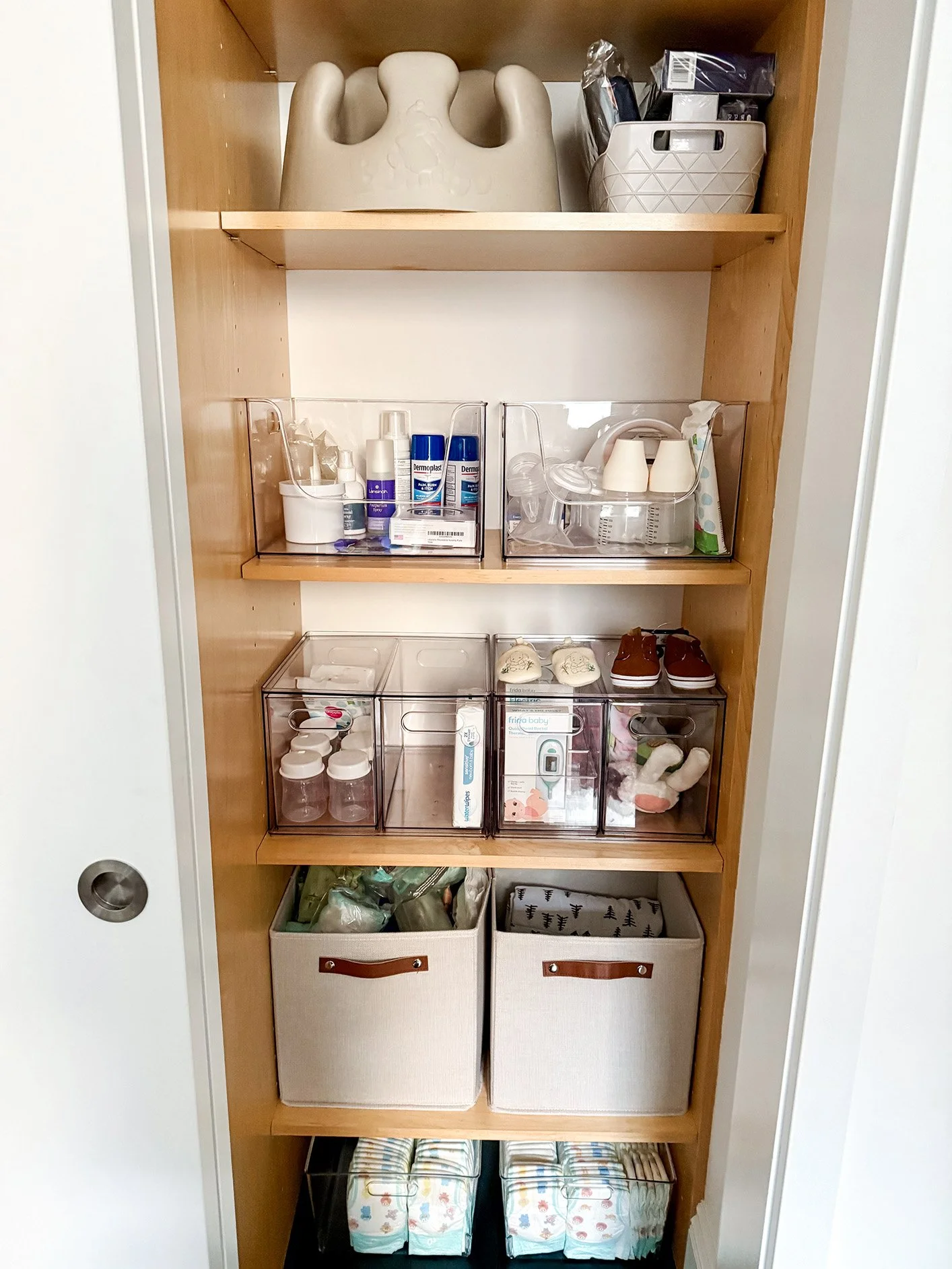 A wooden closet shelf filled with baby supplies including bottles, pacifiers, baby wipes, and diapers organized in clear and fabric storage bins.