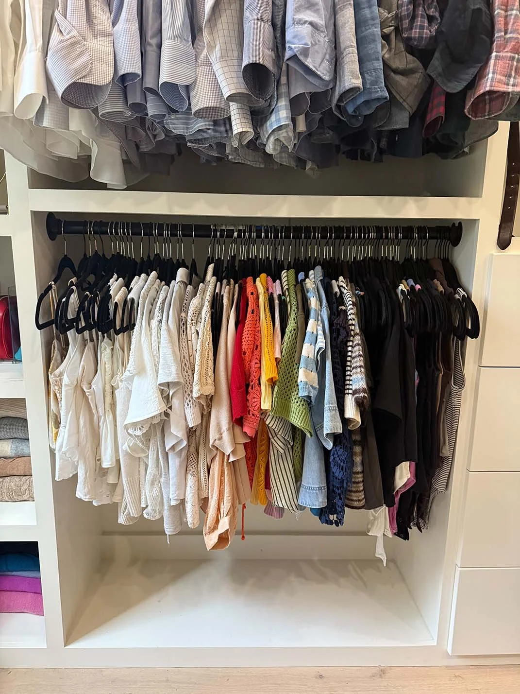 A closet with two shelves of hanging clothes. The top shelf has men's button-up shirts in various shades of blue, gray, and plaid. The bottom shelf has women's blouses in white, cream, red, yellow, green,, and striped patterns, along with black and o