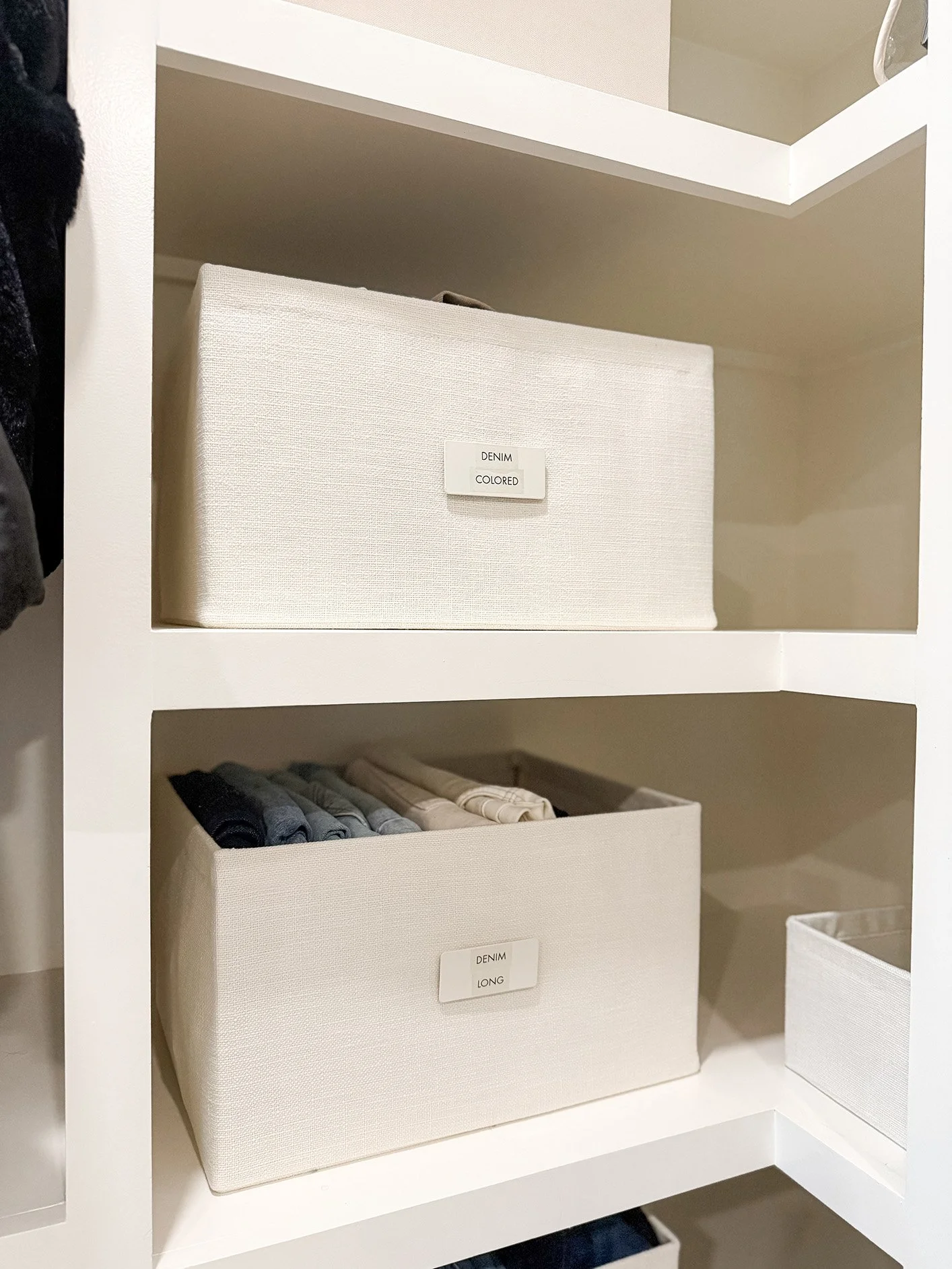 White storage shelves with labeled fabric bins containing folded clothes, labeled 'DENIM COLORED' and 'DENIM LONG'.