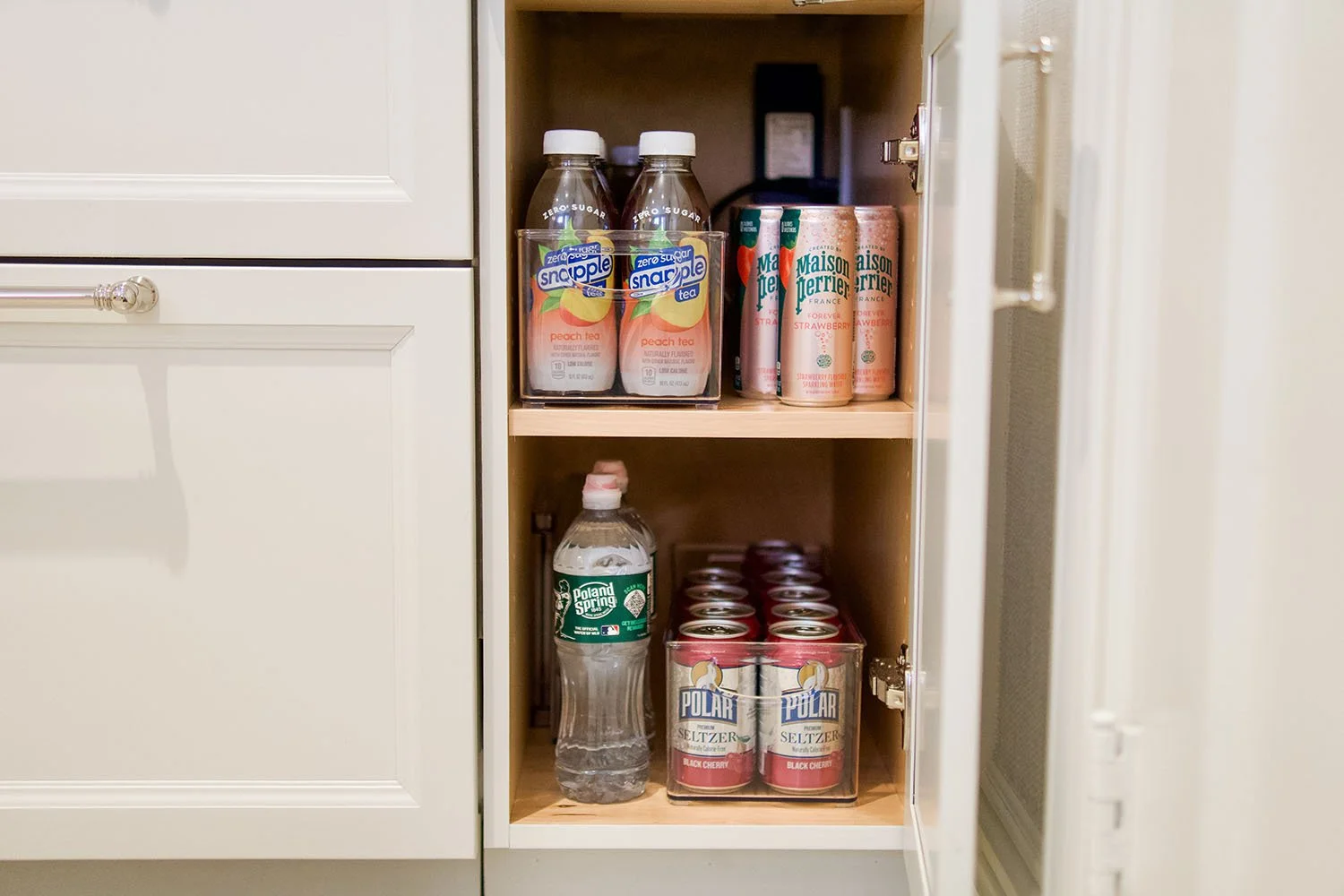 Open built-in kitchen cabinet with two shelves containing beverages: peach zero sugar Snapple tea bottles and Maison Perrier strawberry flavored sparkling water cans on the top shelf, a 16.9 oz Poland Spring water bottle and Polar seltzer cans on the