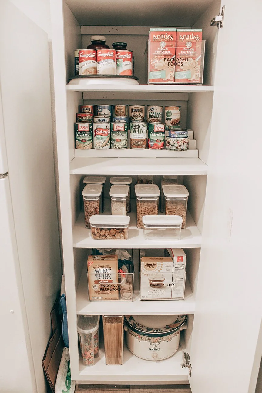 White pantry shelf filled with canned goods, packaged foods, jars, boxes, and a crockpot.