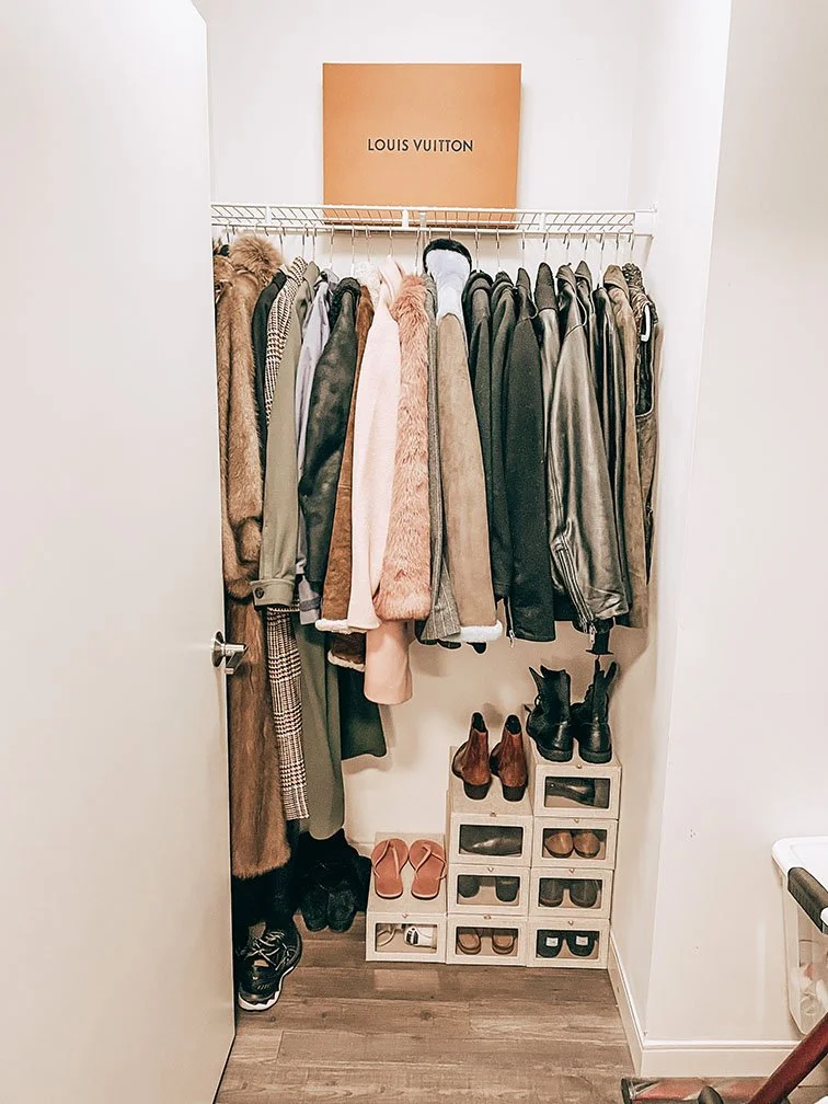A small closet with a clothing rack holding various coats and jackets, and a cardboard Louis Vuitton box on a shelf above. Below the rack, there are shoe cubbies containing multiple pairs of shoes including boots, heels, and sneakers.