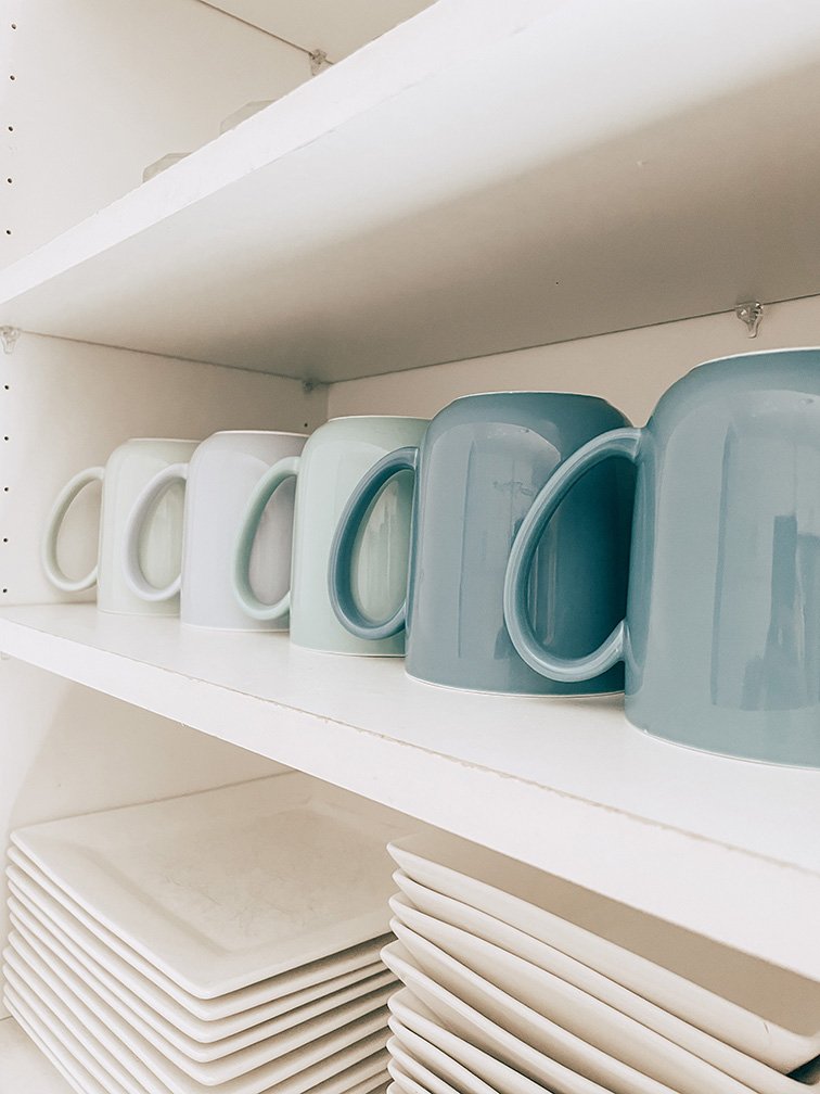 A shelf with six ceramic mugs in shades of white and blue, and a stack of white plates beneath.