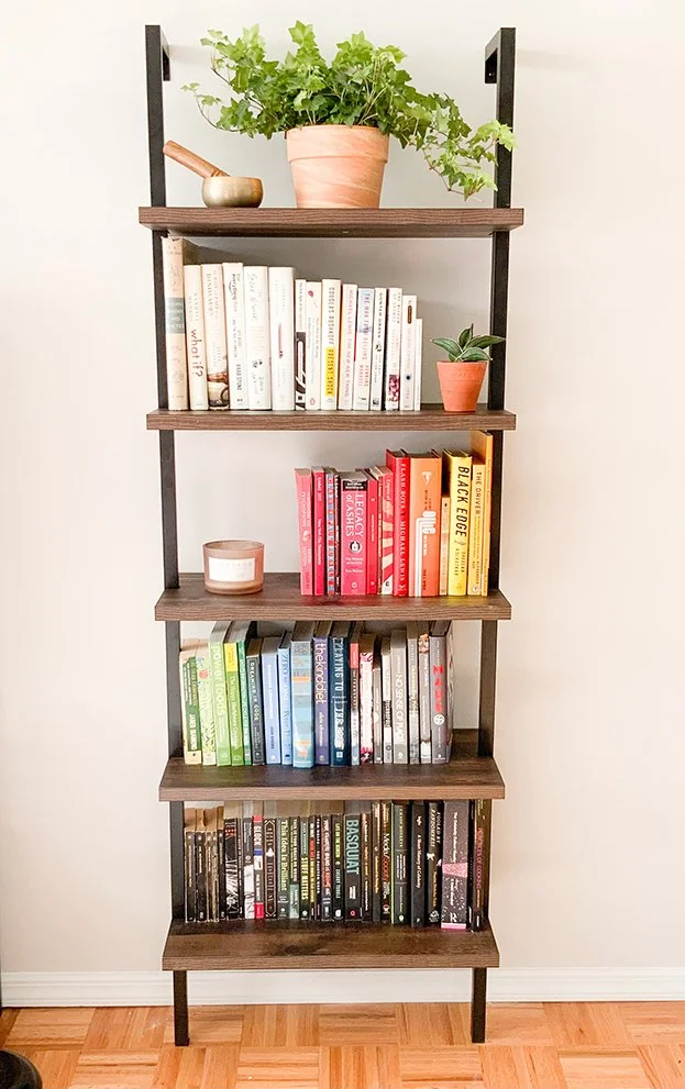 A tall bookshelf with five shelves, decorated with potted plants, books organized by color, and a small candle. The bookshelf is against a white wall, on a hardwood floor.