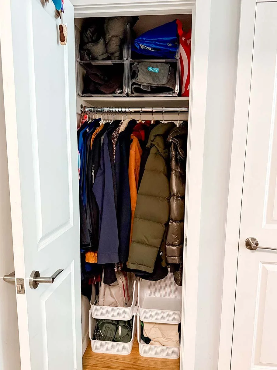 Inside a closet with clothes hanging on a rail, storage bins on shelves above, and baskets on the floor