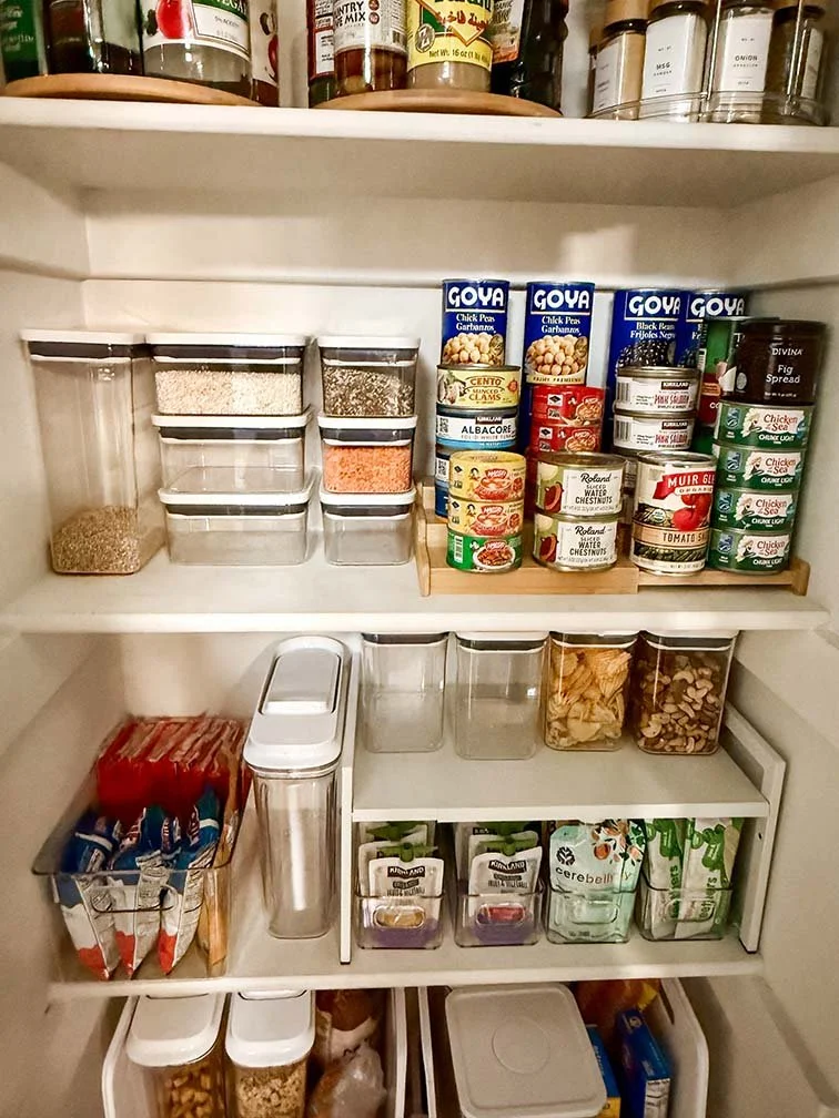 Open refrigerator door showing organized shelves with jars, canned goods, spice containers, snack packs, and drink bottles.