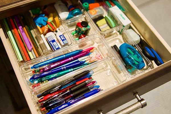 Drawer containing various school supplies including colored pencils, pens, erasers, markers, correction tape, paper clips, small scissors, and adhesive tape.