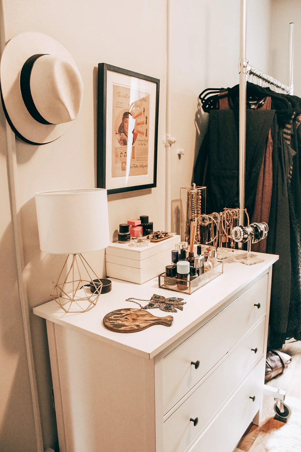 A white dresser with jewelry, nail polish, and a lamp on top, adjacent to a rack of hanging clothes and a framed picture on the wall.