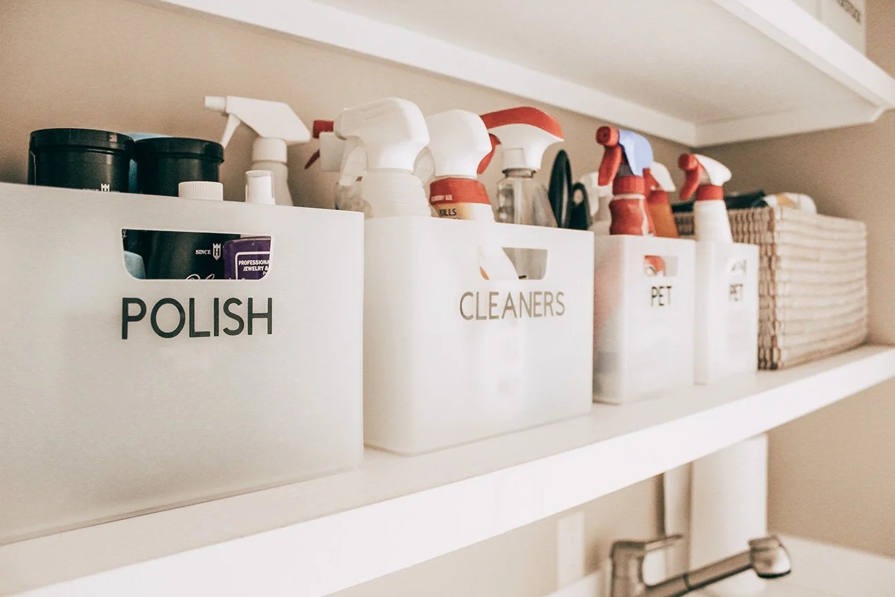 White shelf with storage boxes labeled 'POLISH', 'CLEANERS', and 'PET' filled with spray bottles and cleaning supplies.