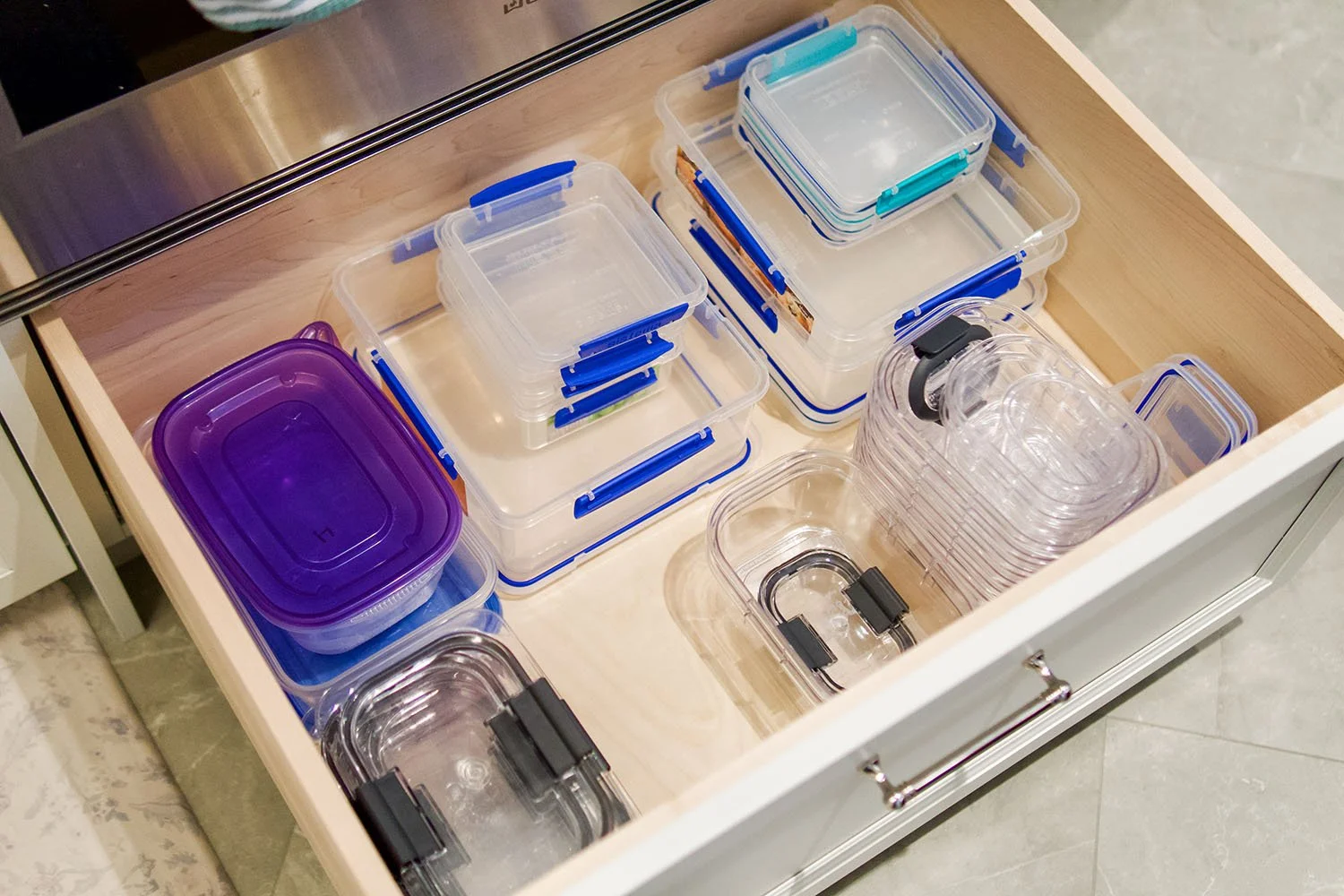 Open kitchen drawer containing various transparent plastic storage containers with blue and black clips, stacked and organized.