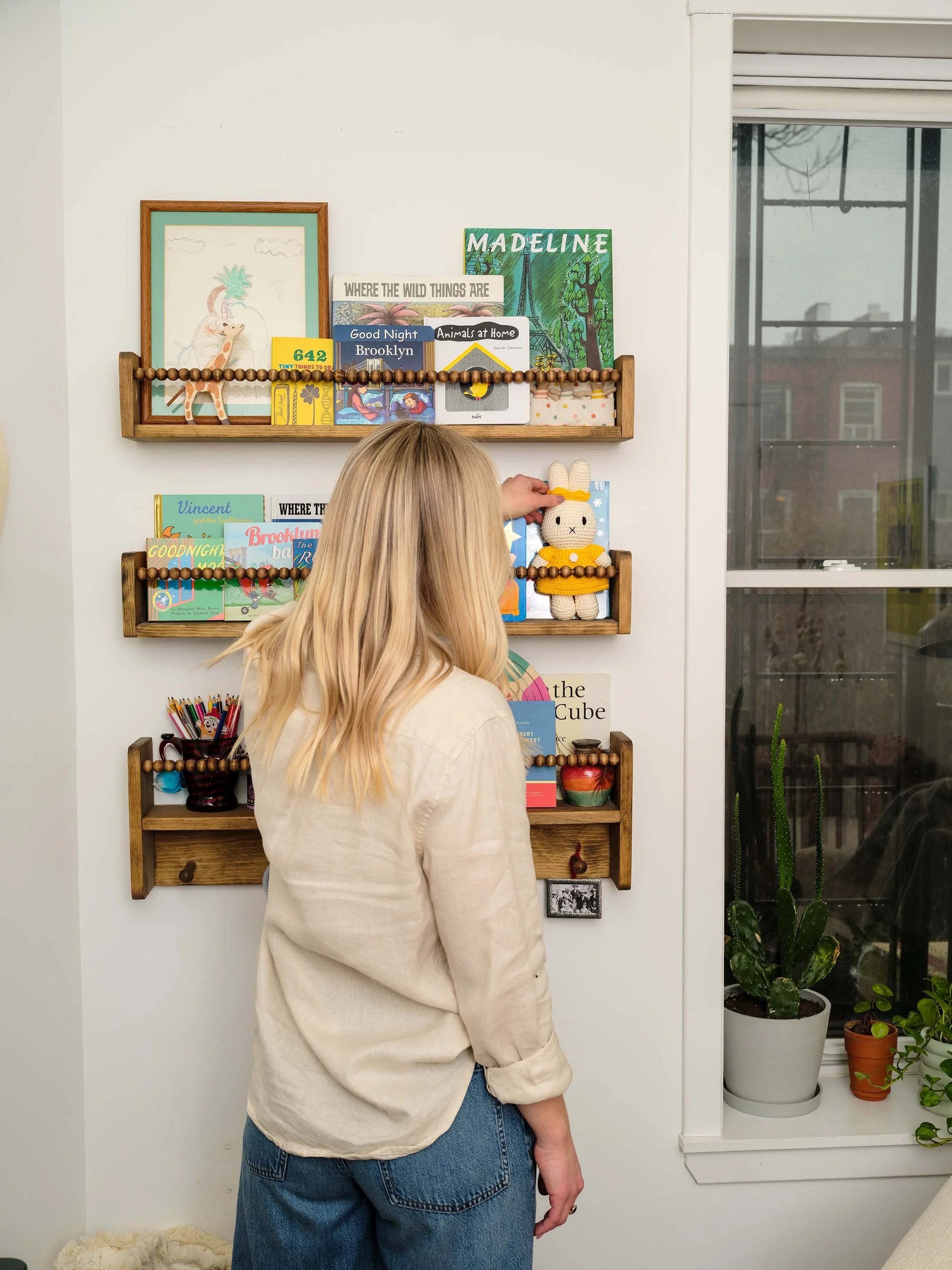 Person with blonde hair wearing a beige jacket and blue jeans, standing in front of wall-mounted shelves filled with children's books, plush toys, and decorative items next to a window with plants outside.