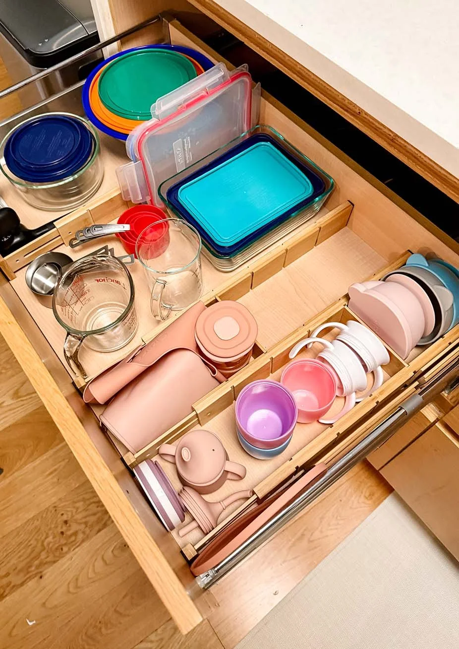 Open kitchen drawer containing various glass and plastic containers, cups, mugs, and kitchen utensils.