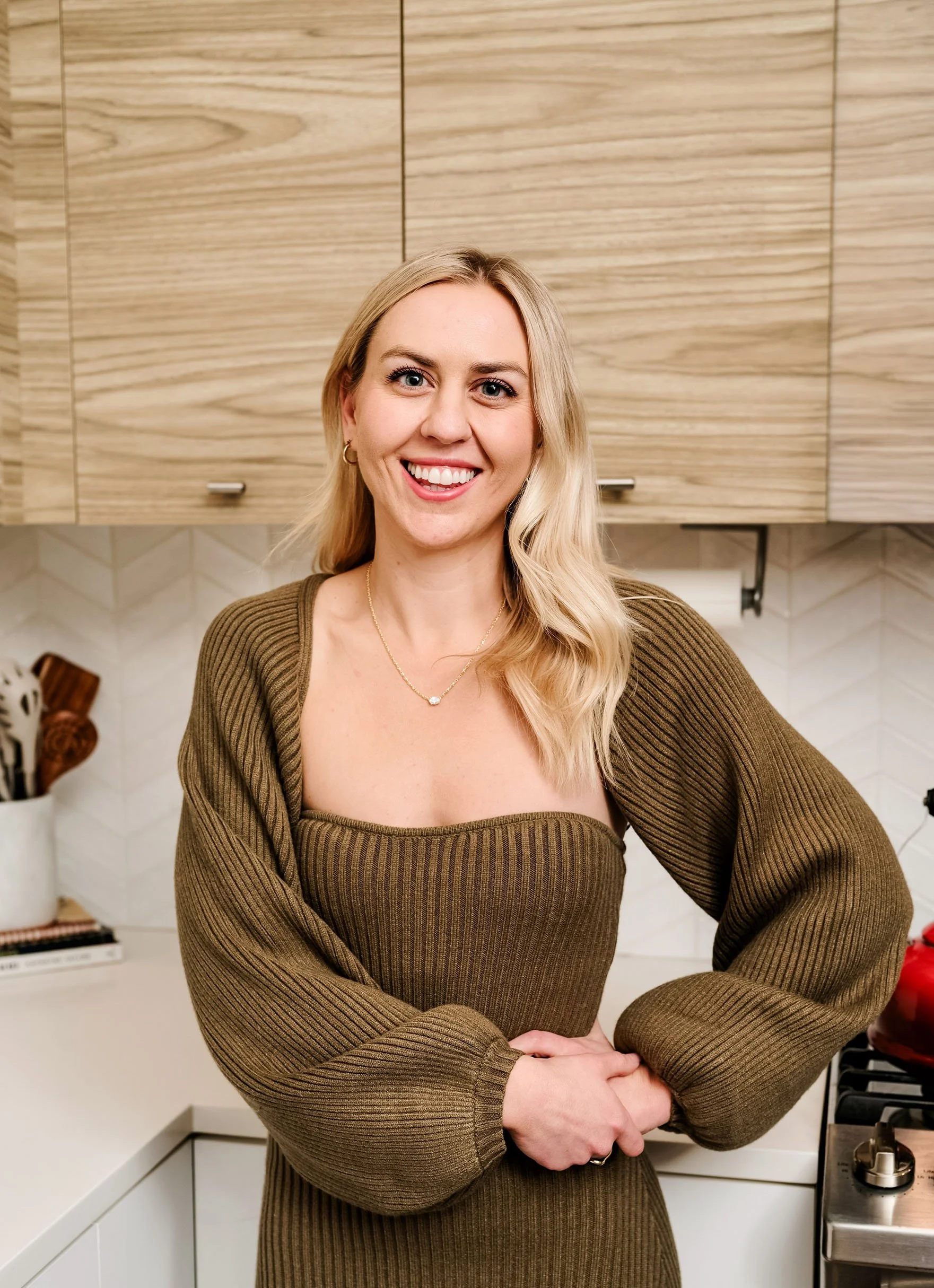 A woman with blonde hair smiling in a kitchen with wooden cabinets and a white backsplash.