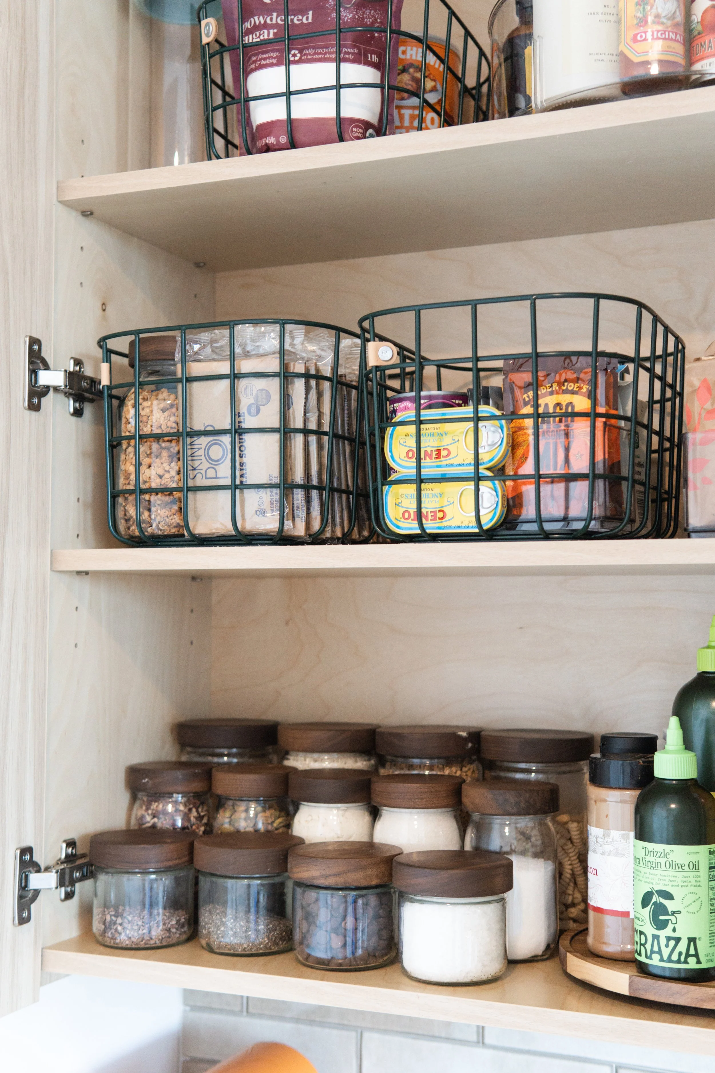 A kitchen cabinet with three wooden shelves, holding various pantry items including spices in glass jars, canned foods, and cooking oils, with metal baskets on the upper shelf.