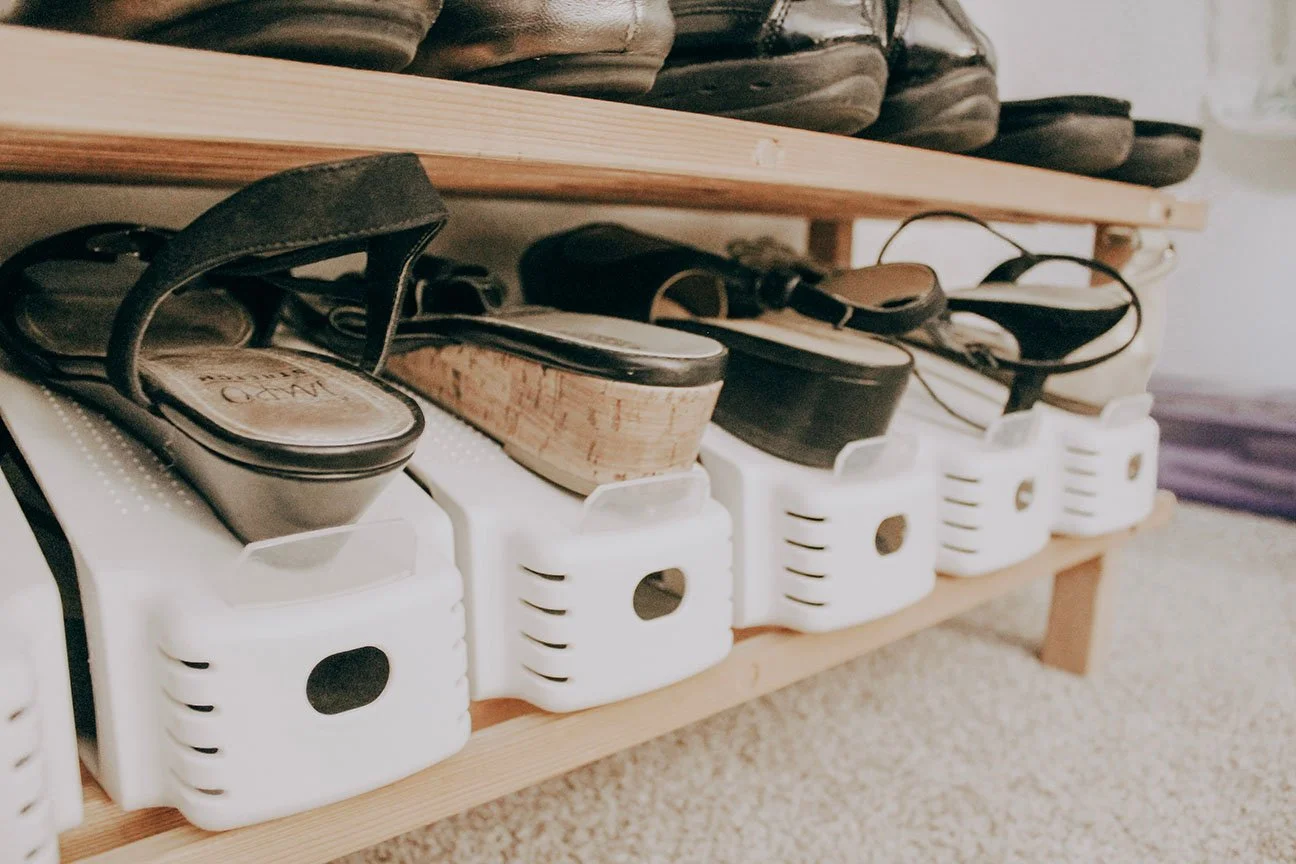 Close-up of a row of footwear on white shoe racks, including black sandals and wedge shoes, with a wooden shelf above.