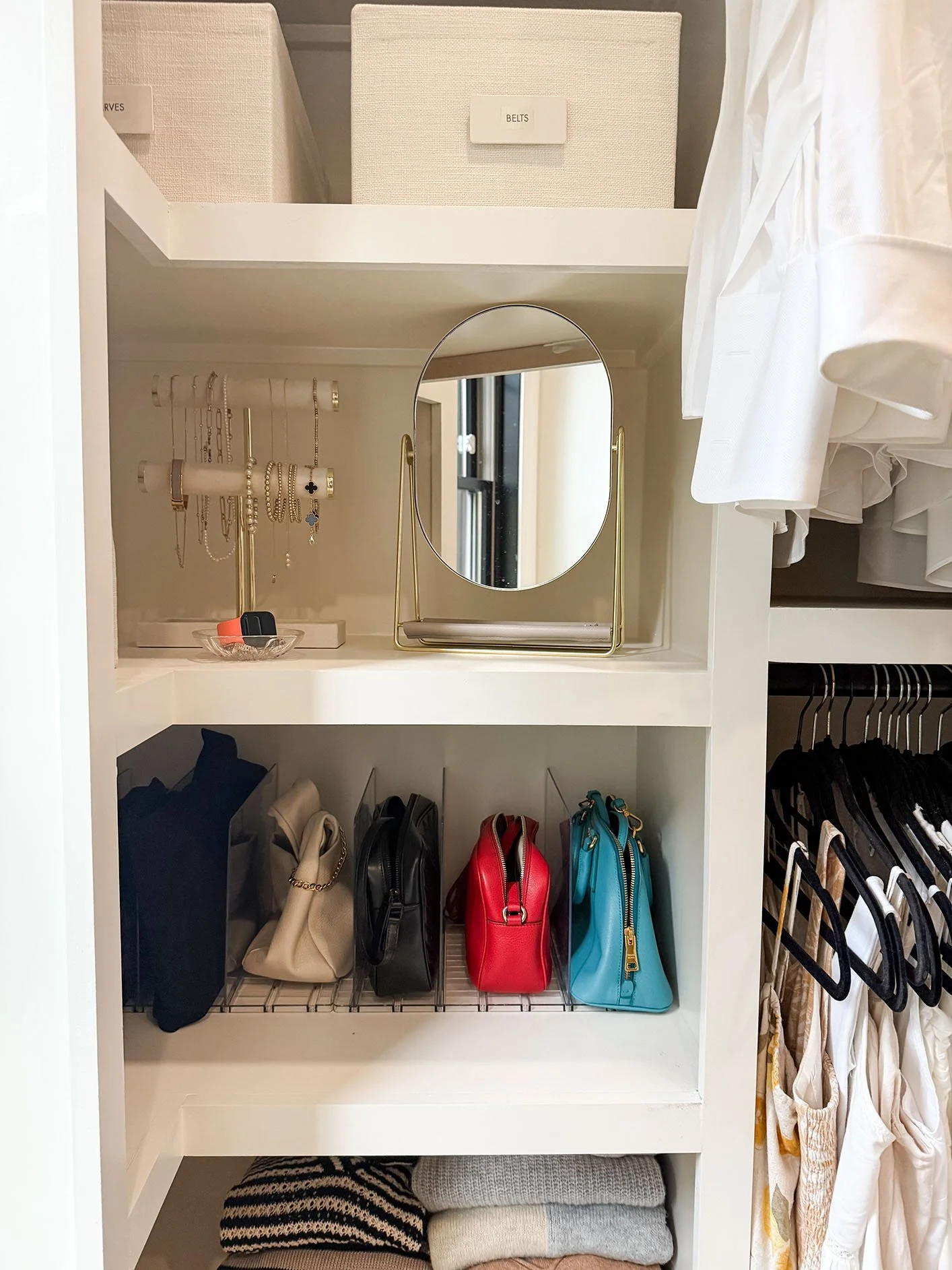 A closet organizer with shelves and hanging space. The upper shelf has storage boxes labeled 'BELTS' and 'PURSES.' The middle shelf has a mirror and jewelry stand with necklaces and bracelets. The bottom shelf has handbags in different colors. On the