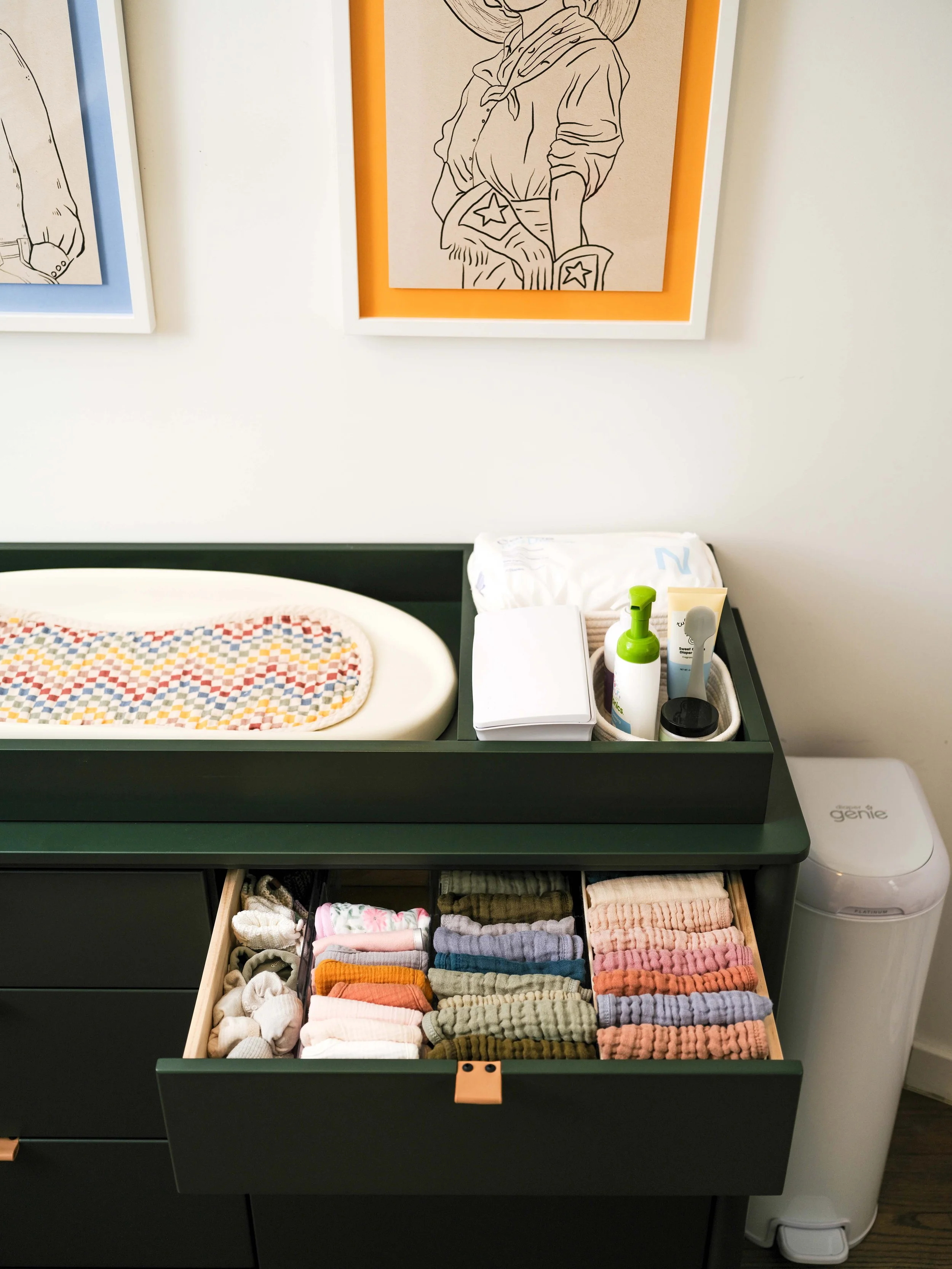 A changing table with a colorful cloth on it, a drawer filled with neatly folded socks of various colors, and a container holding toiletries including lotion, a toothbrush, and a baby wipe. There is a framed artwork on the wall and a white device lab