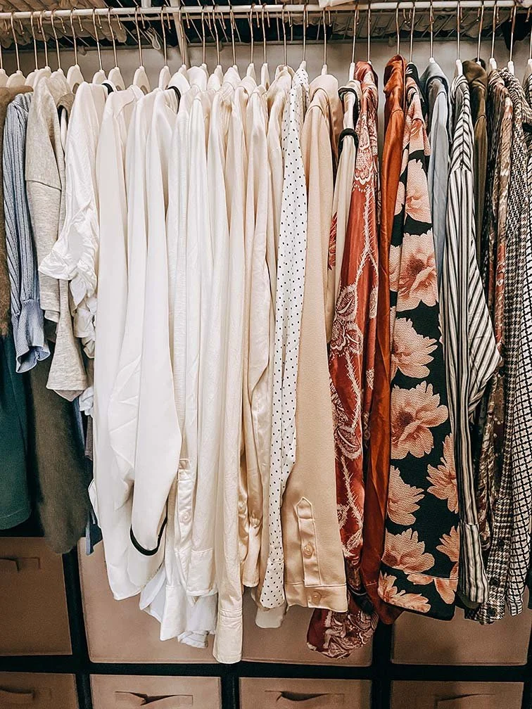 Clothes hanging on a rack, including white, beige, patterned, floral, striped, and checkered shirts or dresses.